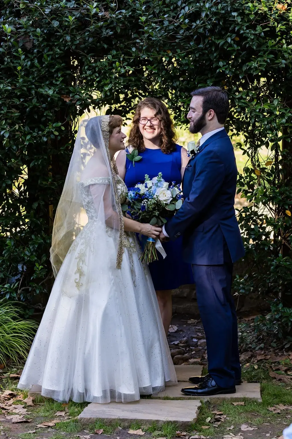 Jessica and Hayden face each other at the garden altar under a leafy arch as an attendant smiles