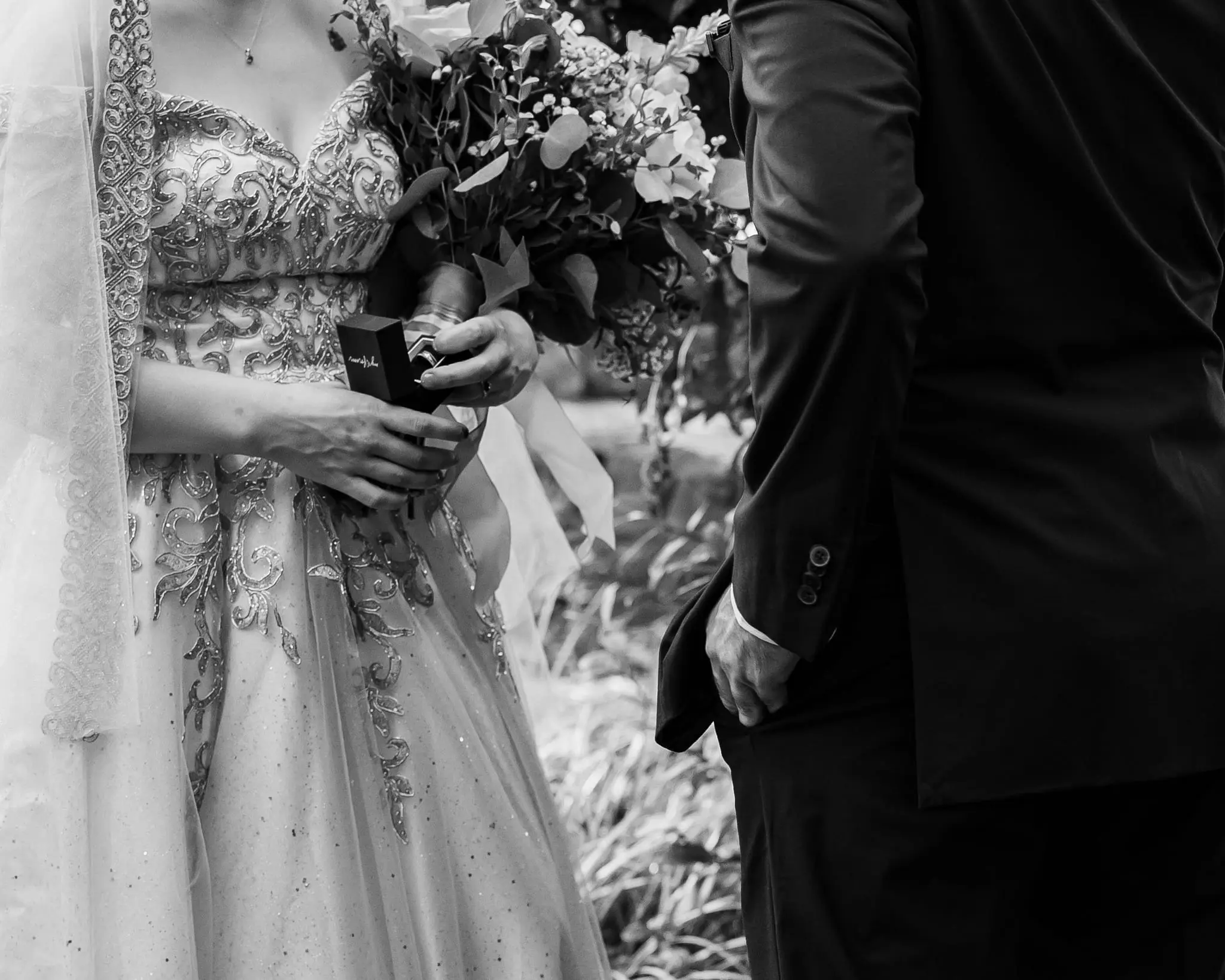 Black and white detail of the bride's gold-embroidered gown and bouquet as she holds a ring box