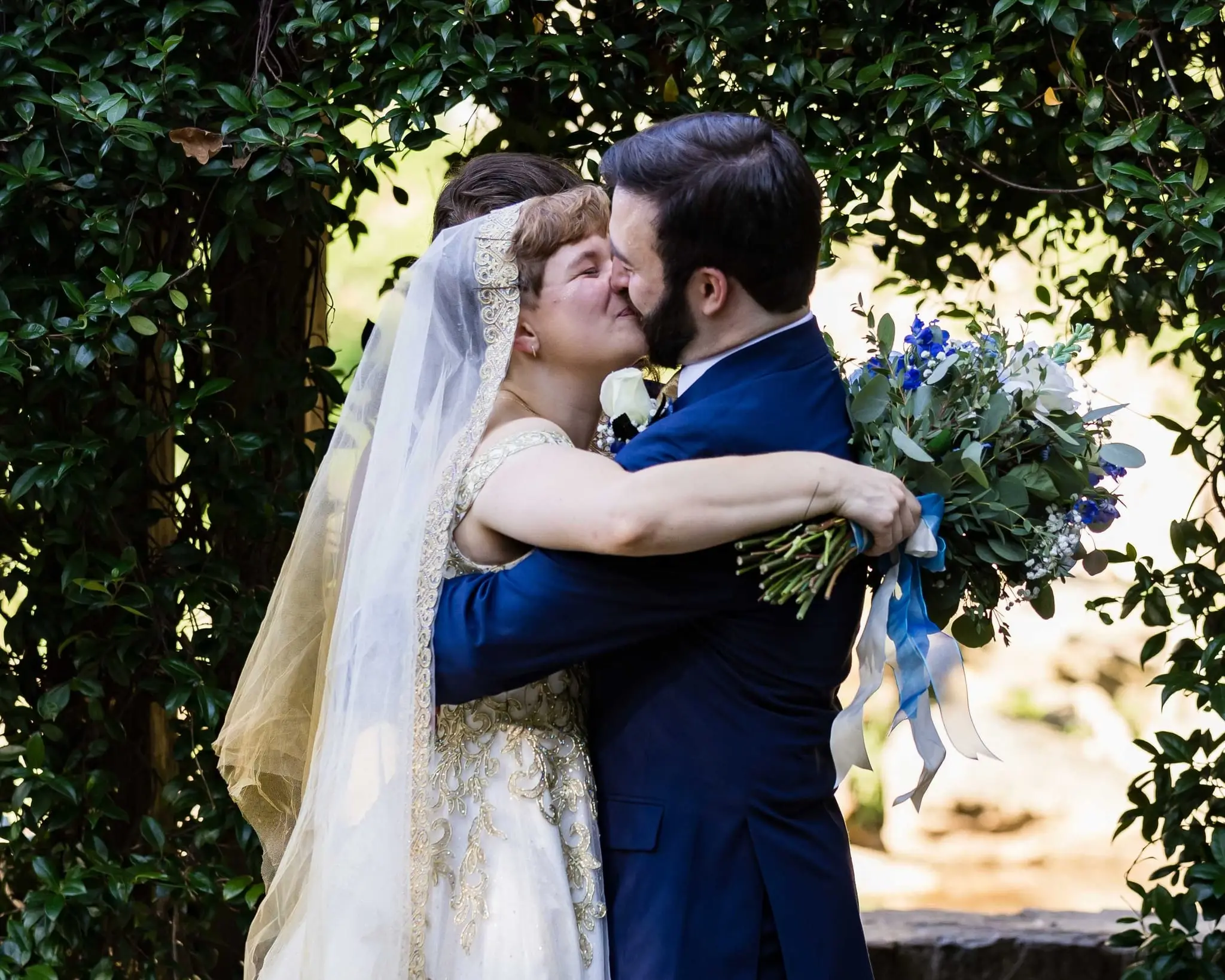 Newlyweds kiss in an embrace beneath leafy greenery, the bride's bouquet in hand