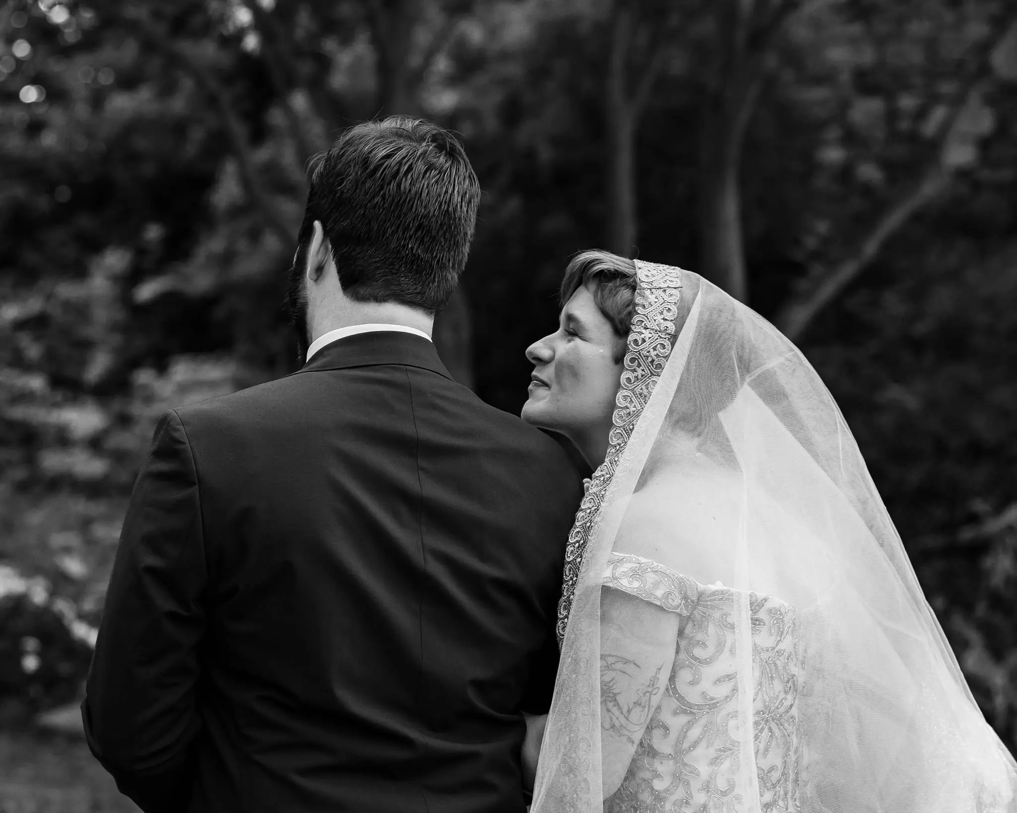 Black and white of the bride in a lace-edged veil resting her head against the groom's shoulder
