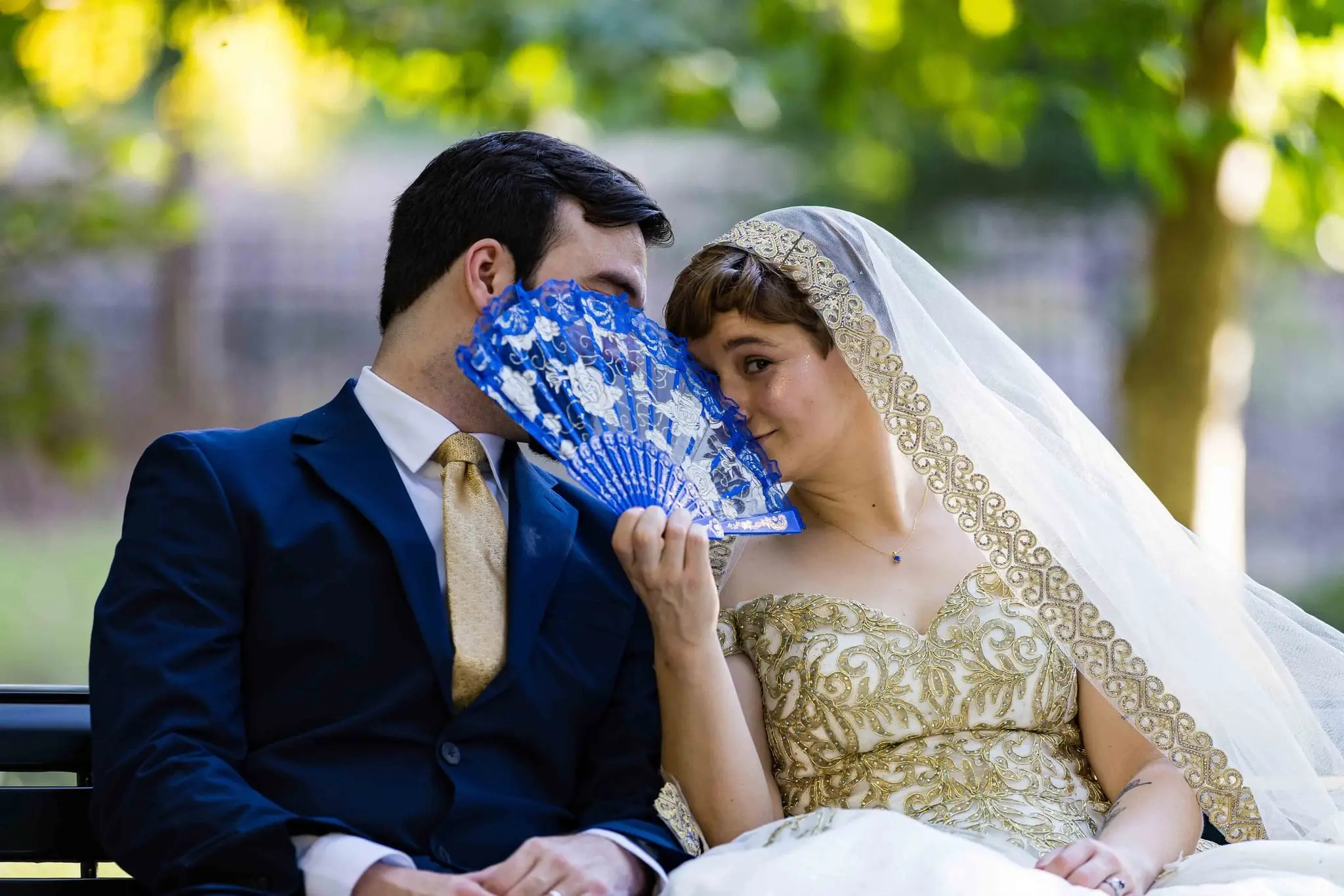 Groom and bride nuzzling behind a blue folding fan on a park bench