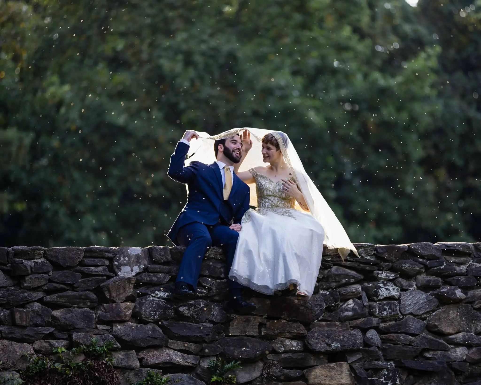 Couple sitting on a stone wall sharing a laugh under the bride's veil