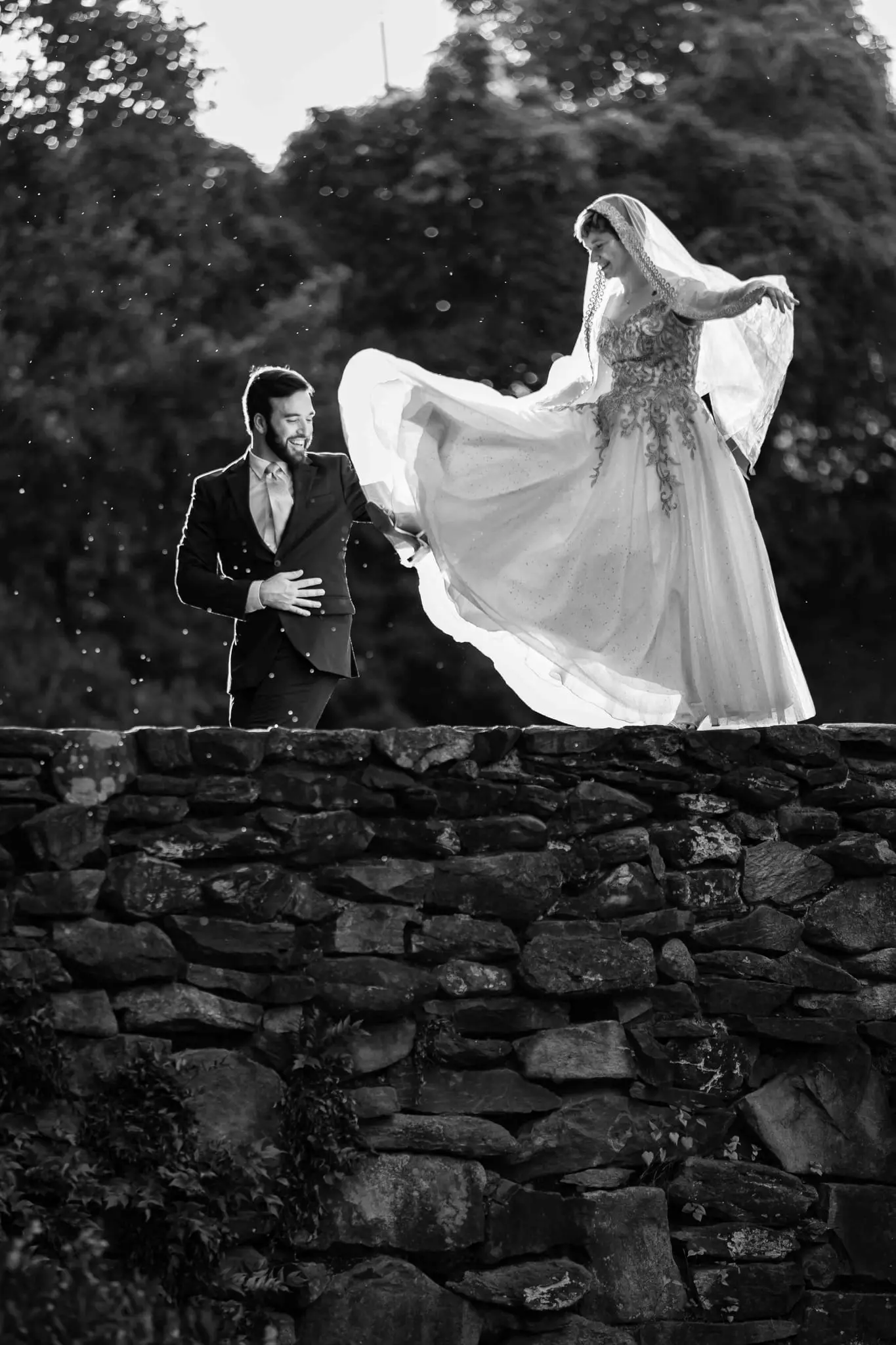 Black and white of the bride flaring her gown atop a stone wall as the groom looks on