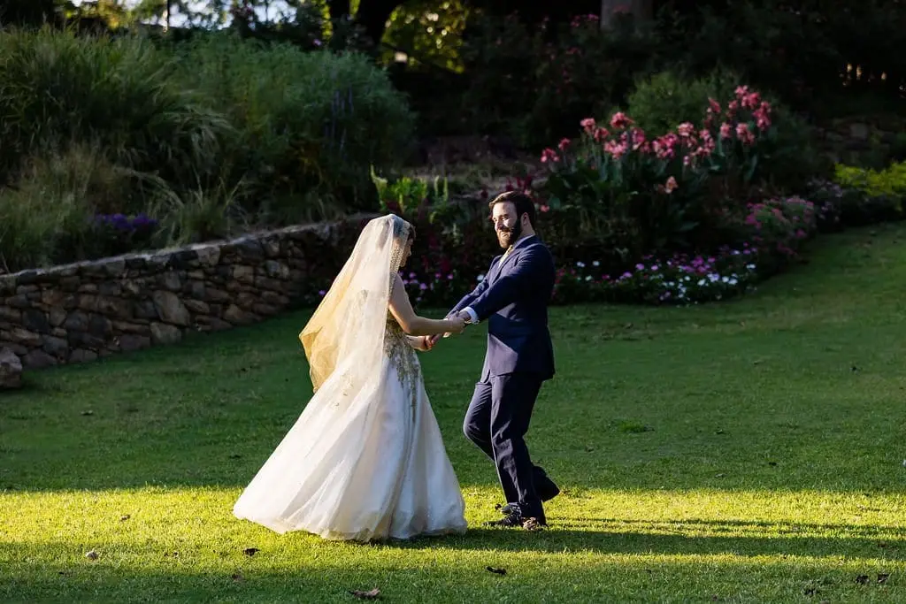 Bride in a gold-trimmed veil dancing with the groom on a sunlit lawn beside a stone wall