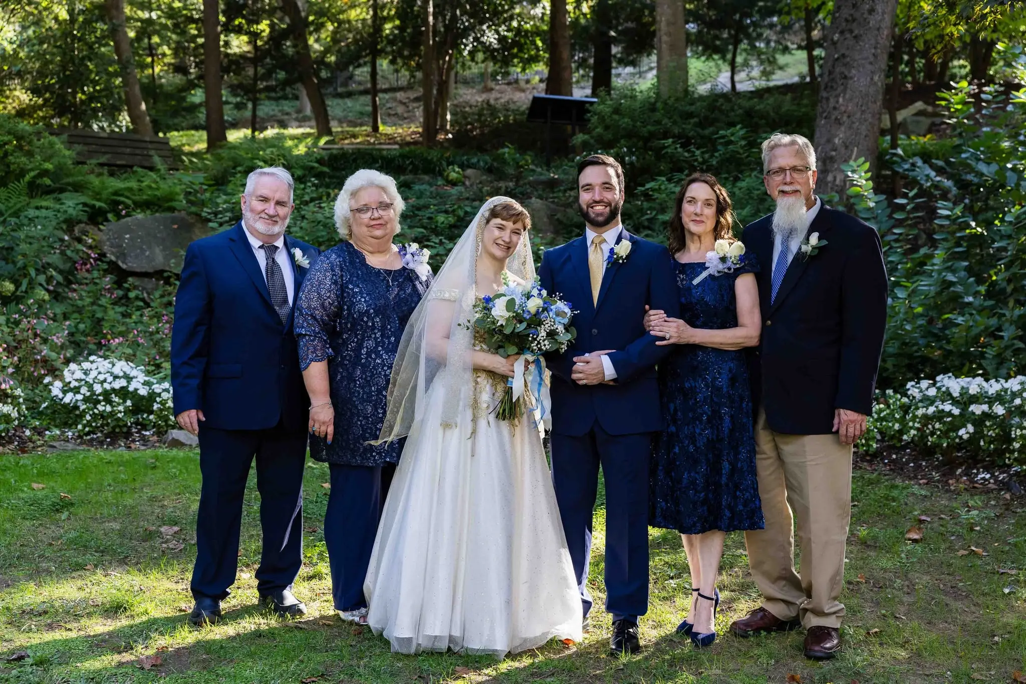 Bride and groom posed with four older family members on a green lawn bordered by white flowers