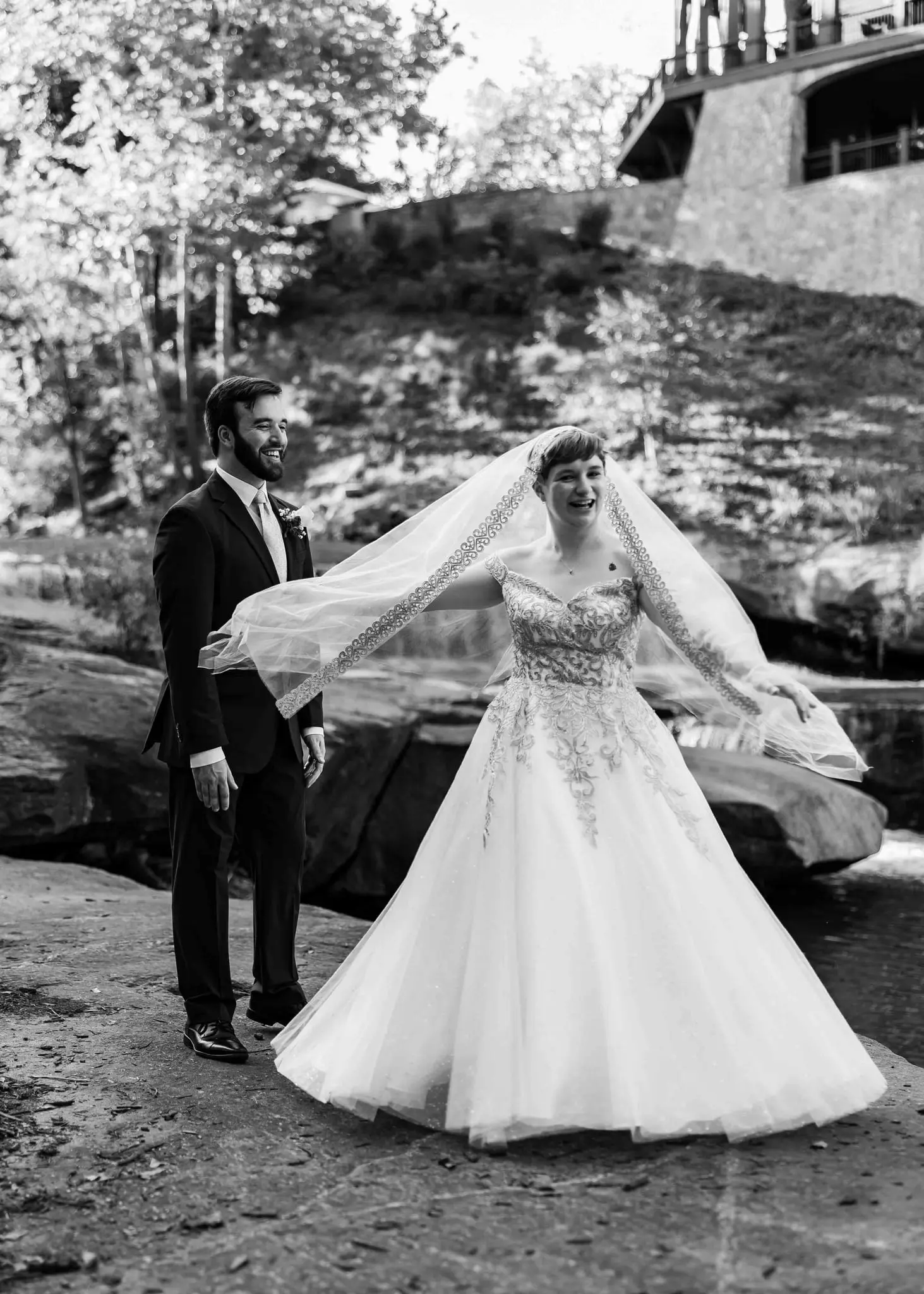 Black and white of the bride twirling her veil on the river rocks as the groom looks on
