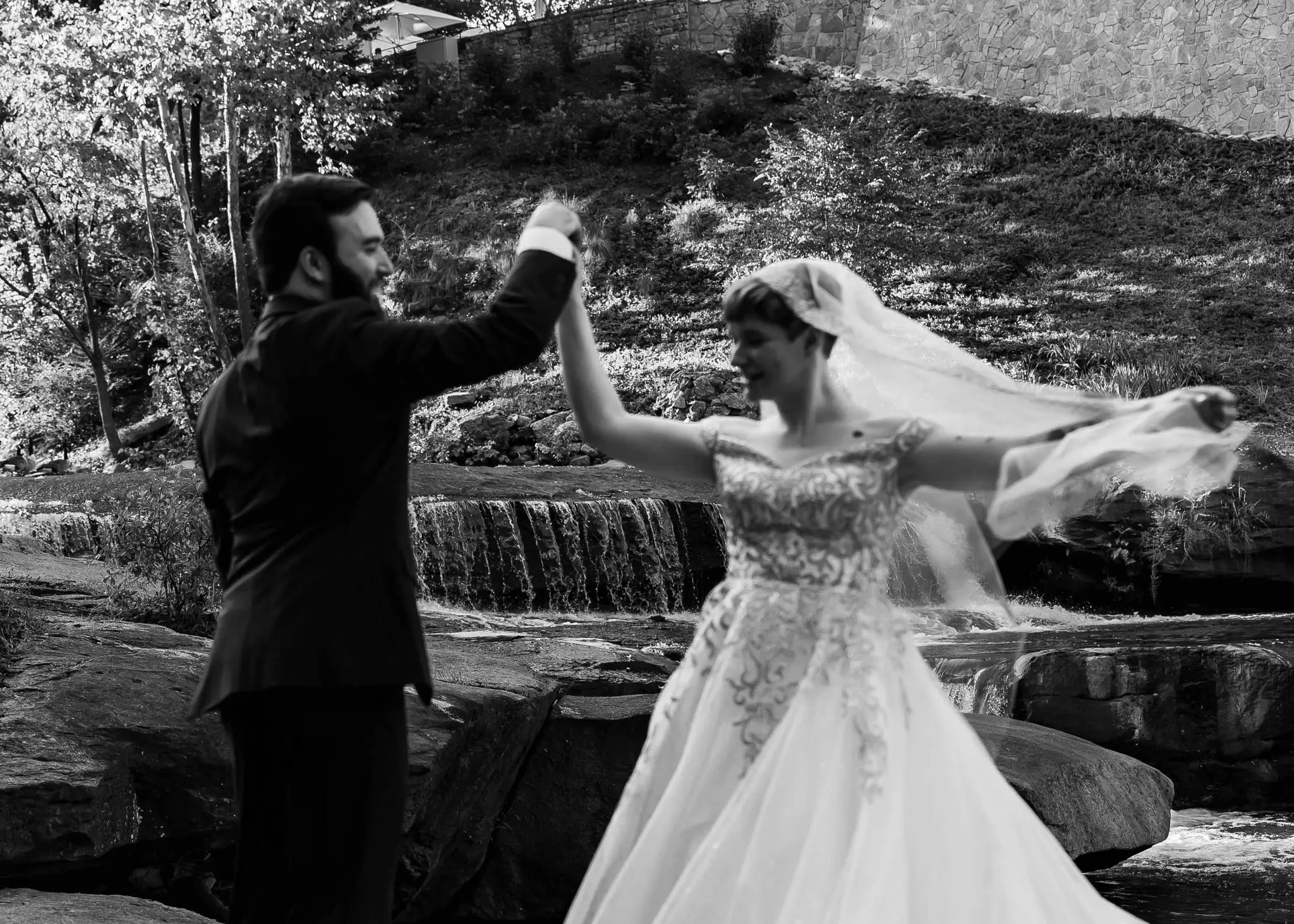 Black and white of the groom twirling the bride beside a waterfall, her veil flying