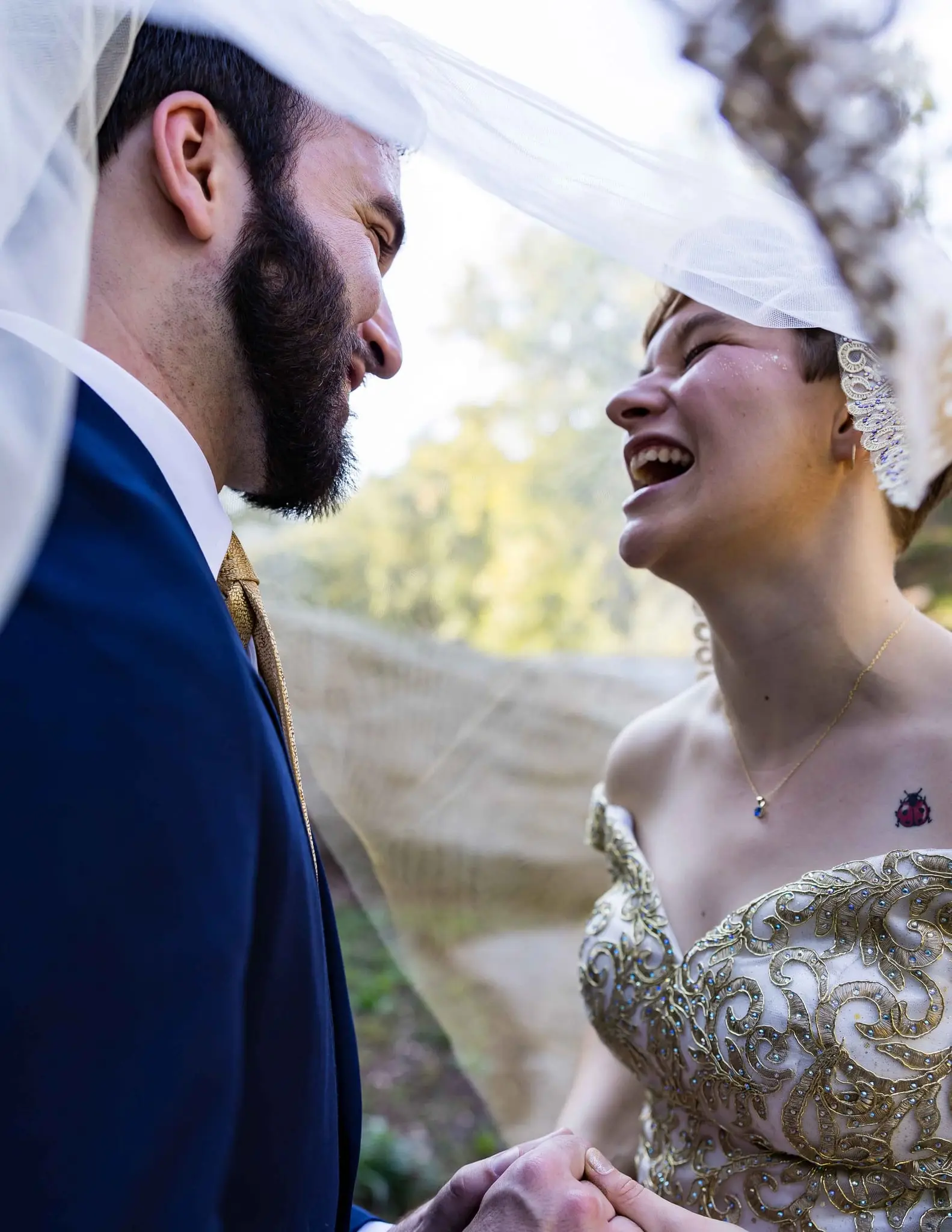 Groom and bride laughing face to face beneath her veil while holding hands