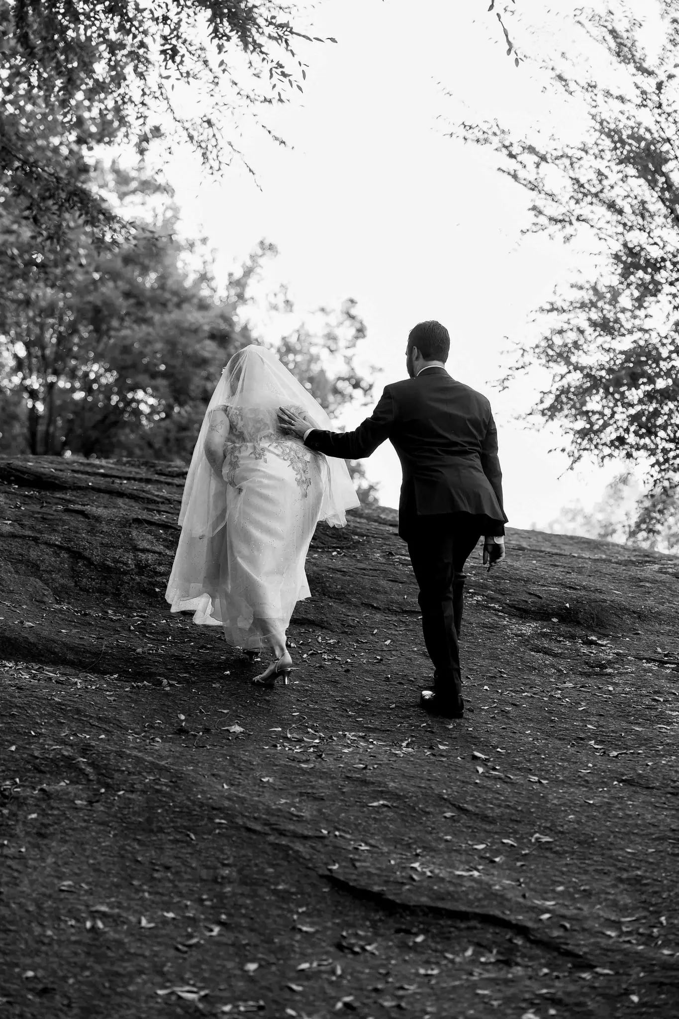 Black and white of the groom helping the bride up a sloping rock face
