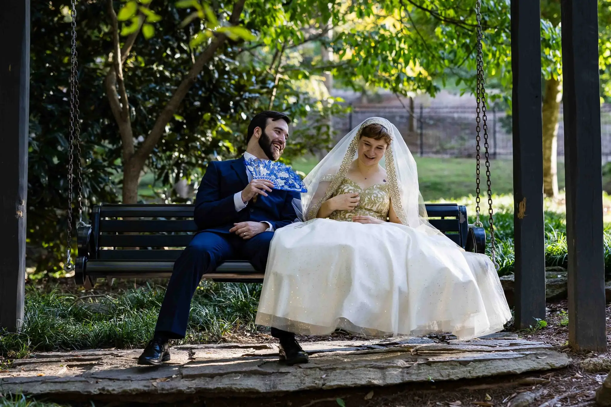 Jessica and Hayden wedding at Grand Bohemian Lodge Greenville, SC — the couple sitting on a bench under a pavilion, the groom holding a blue fan