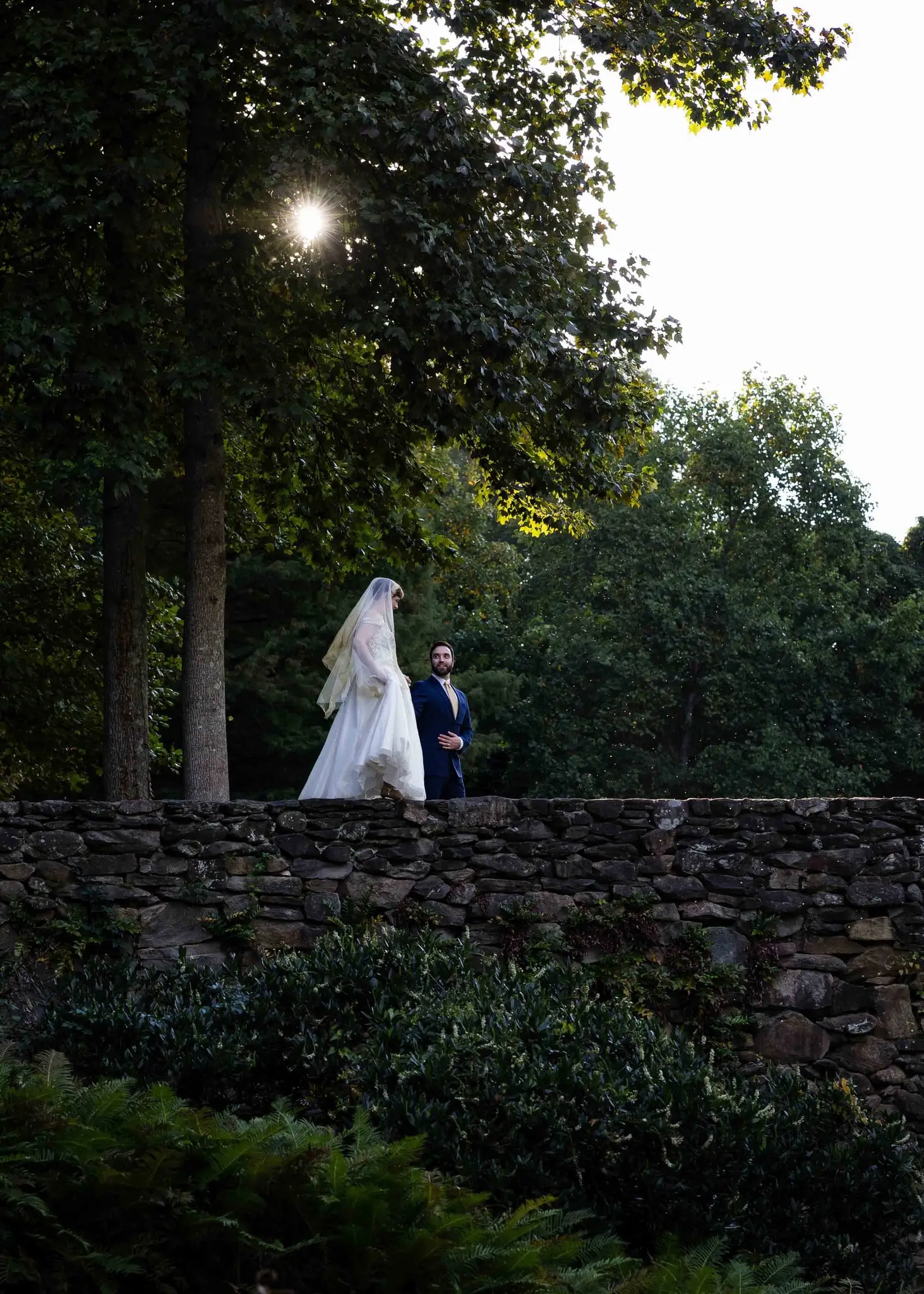 Bride and groom small in the frame along a stone wall beneath sunlit trees