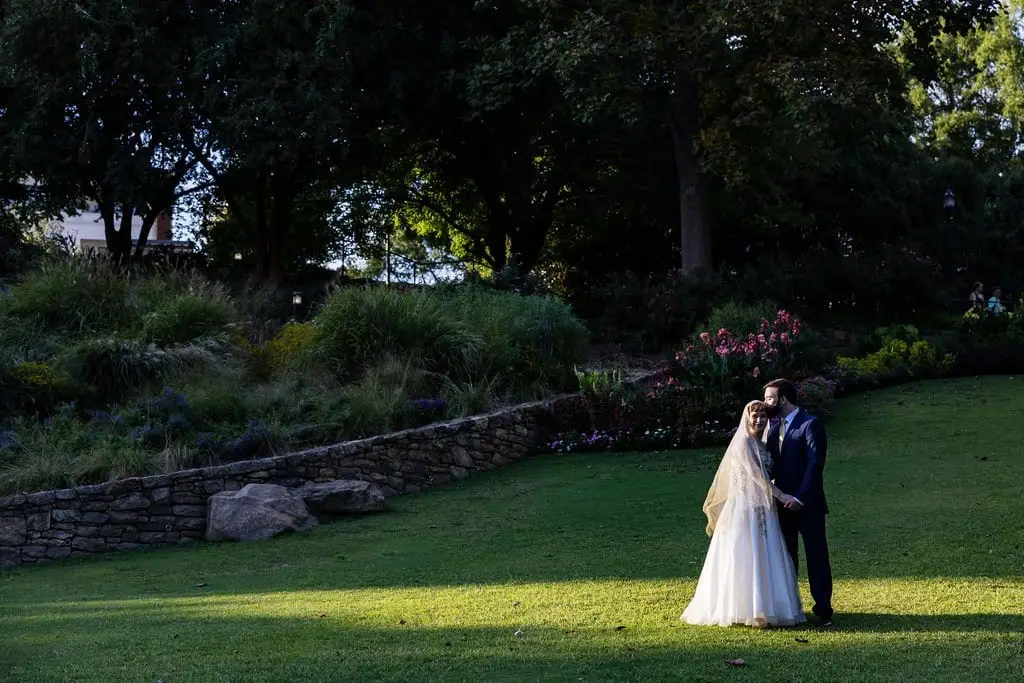 Couple embracing in the distance on a sunlit lawn with a stone retaining wall and flower beds
