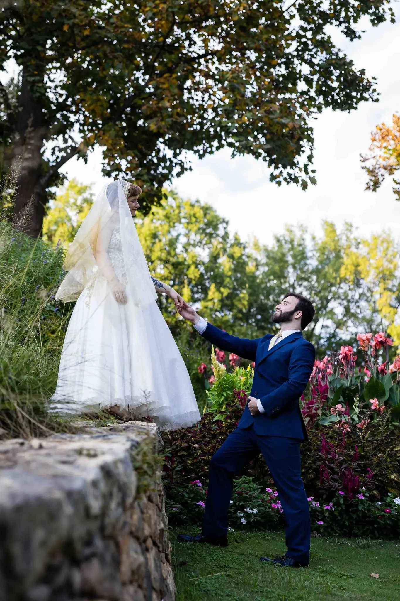 Bride standing atop a stone garden wall as the groom reaches up to take her hand beside pink flowers