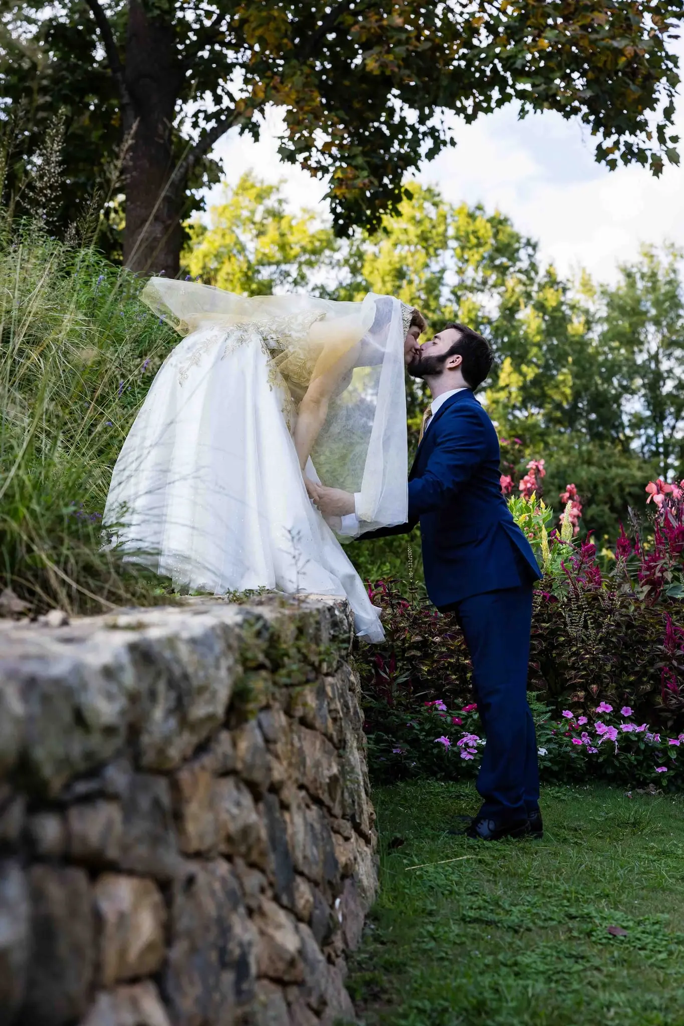 Groom leaning in for a kiss as the bride bends down from a stone wall, her veil falling forward