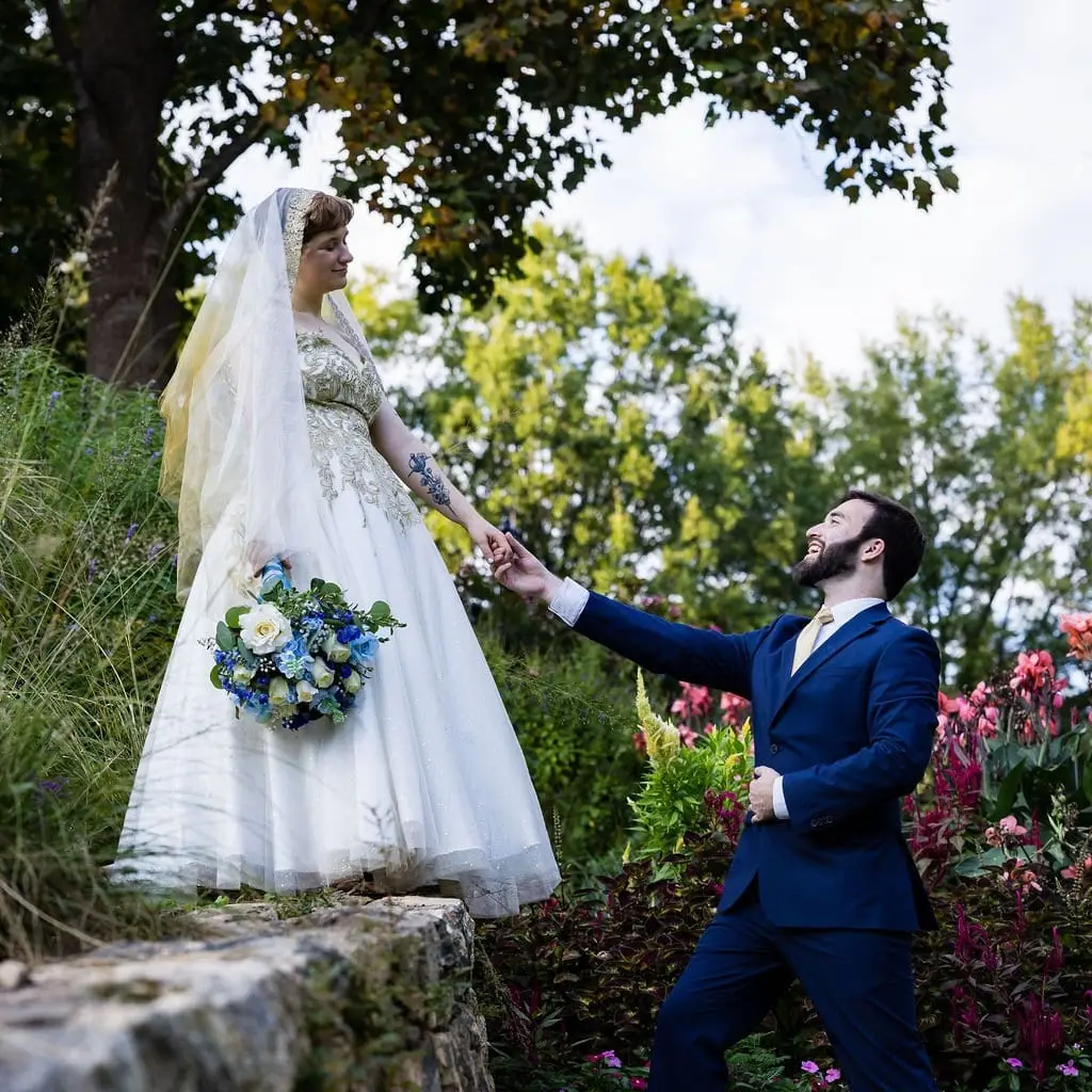 Hayden in a navy suit reaching up to take Jessica's hand as she stands on a stone wall above flower beds