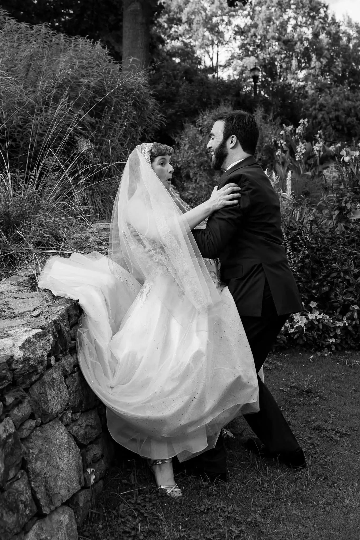 Black and white of the bride leaning into the groom beside a stone garden wall, her skirt billowing