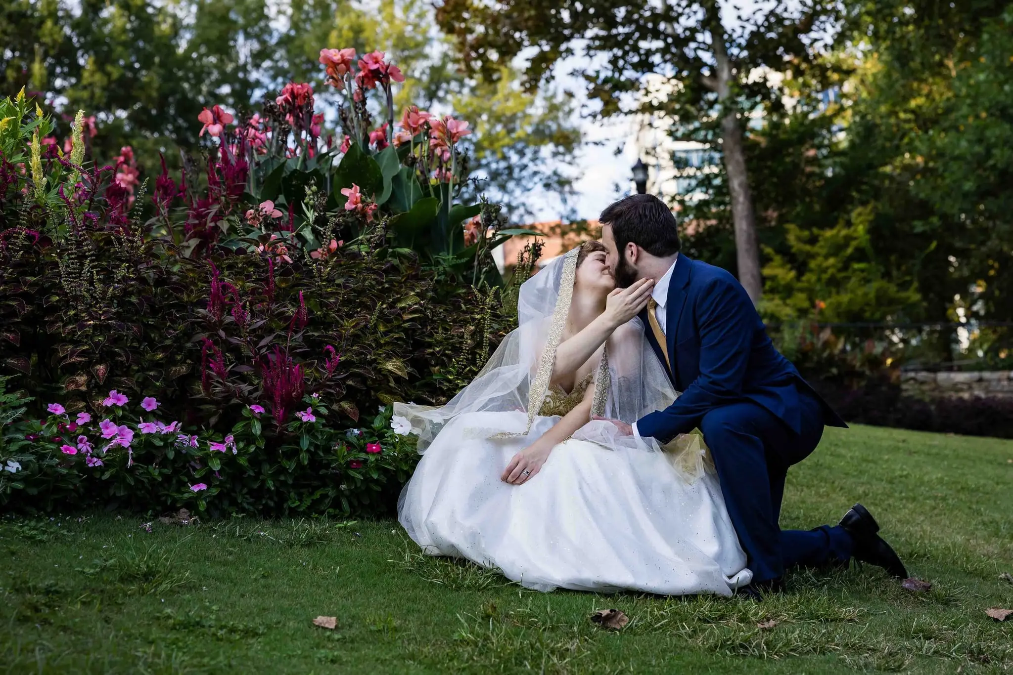 Couple kissing on the lawn, the bride seated in her gown and the groom kneeling among magenta flowers