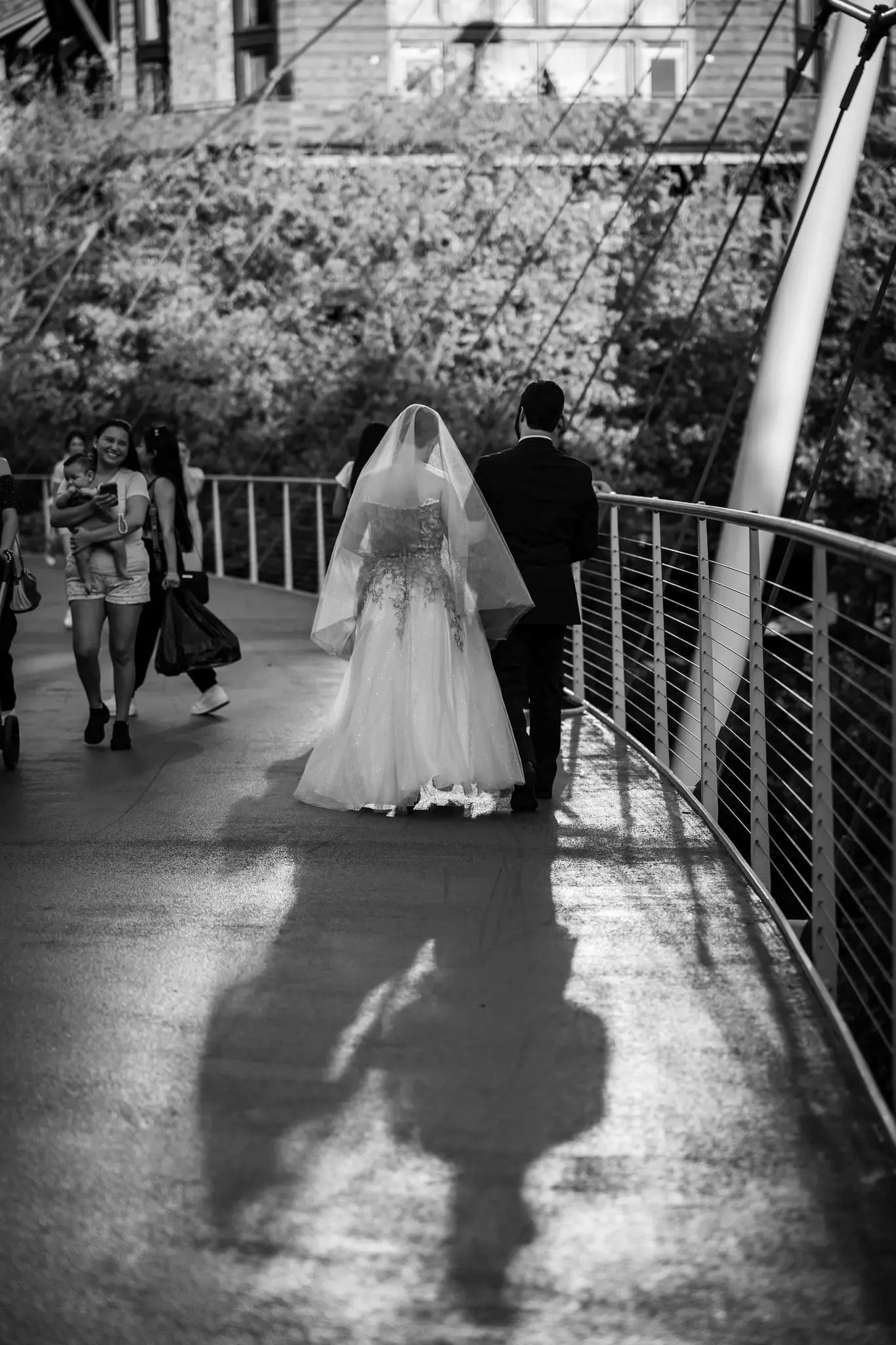 Black and white of the bride and groom walking along a curved pedestrian bridge, their shadows long beside them