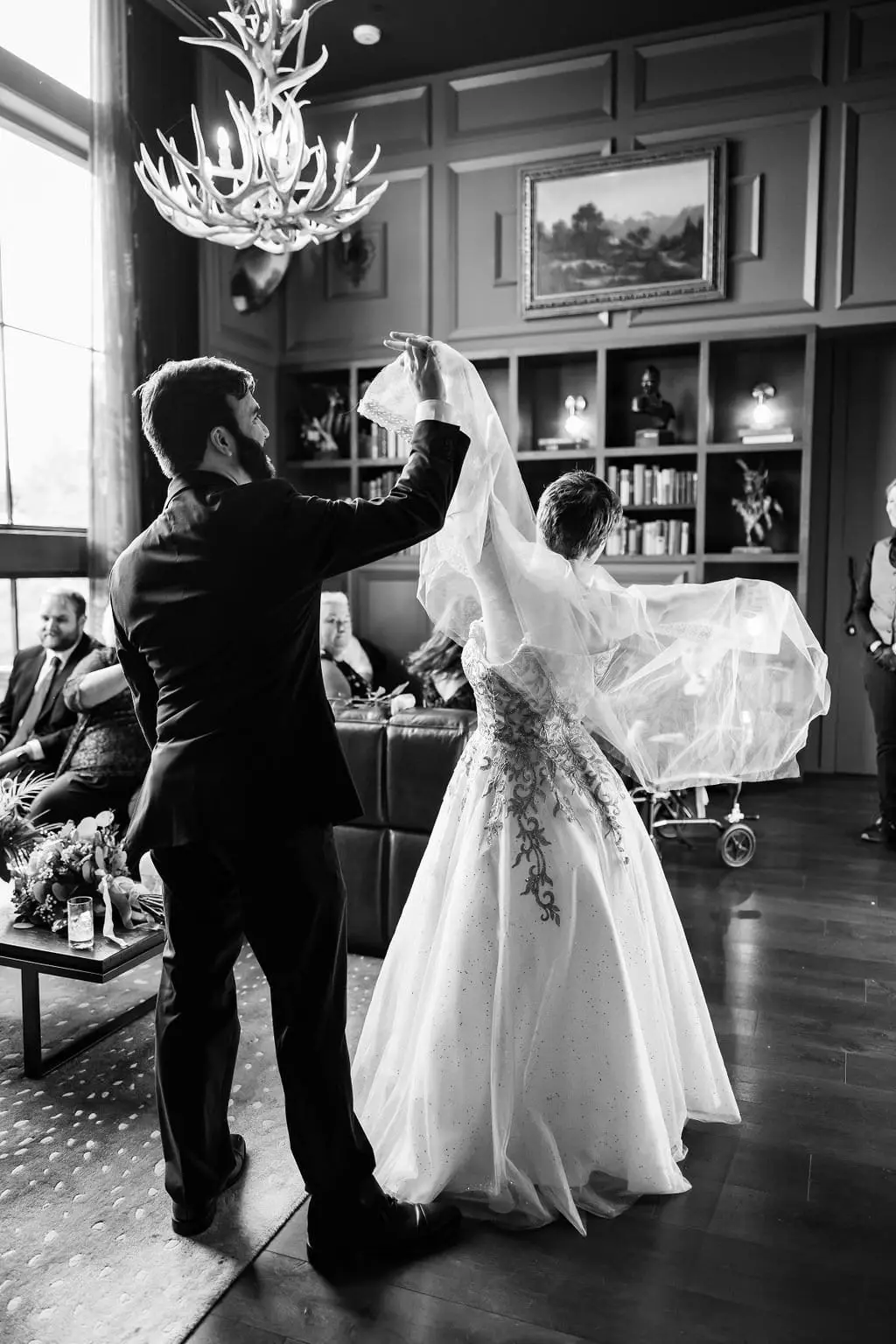 Black and white of Hayden twirling Jessica's veil beneath an antler chandelier in the hotel lounge