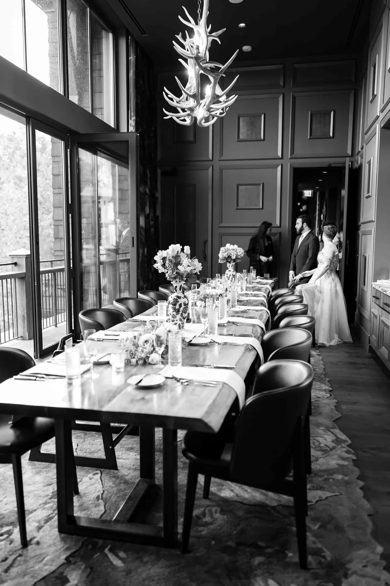 Black and white of the long reception table beneath an antler chandelier as the couple wait at the far end