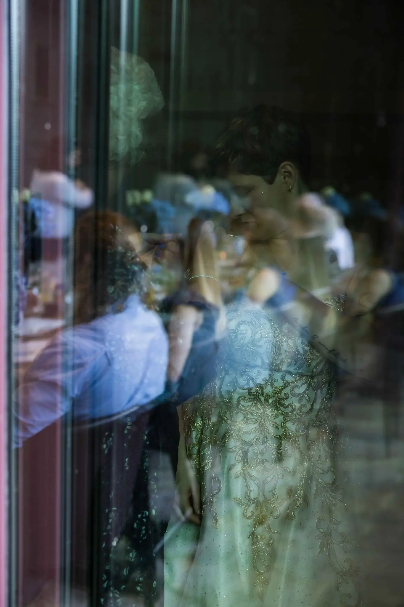 Window reflection layering the bride's gold-beaded gown with blurred guests