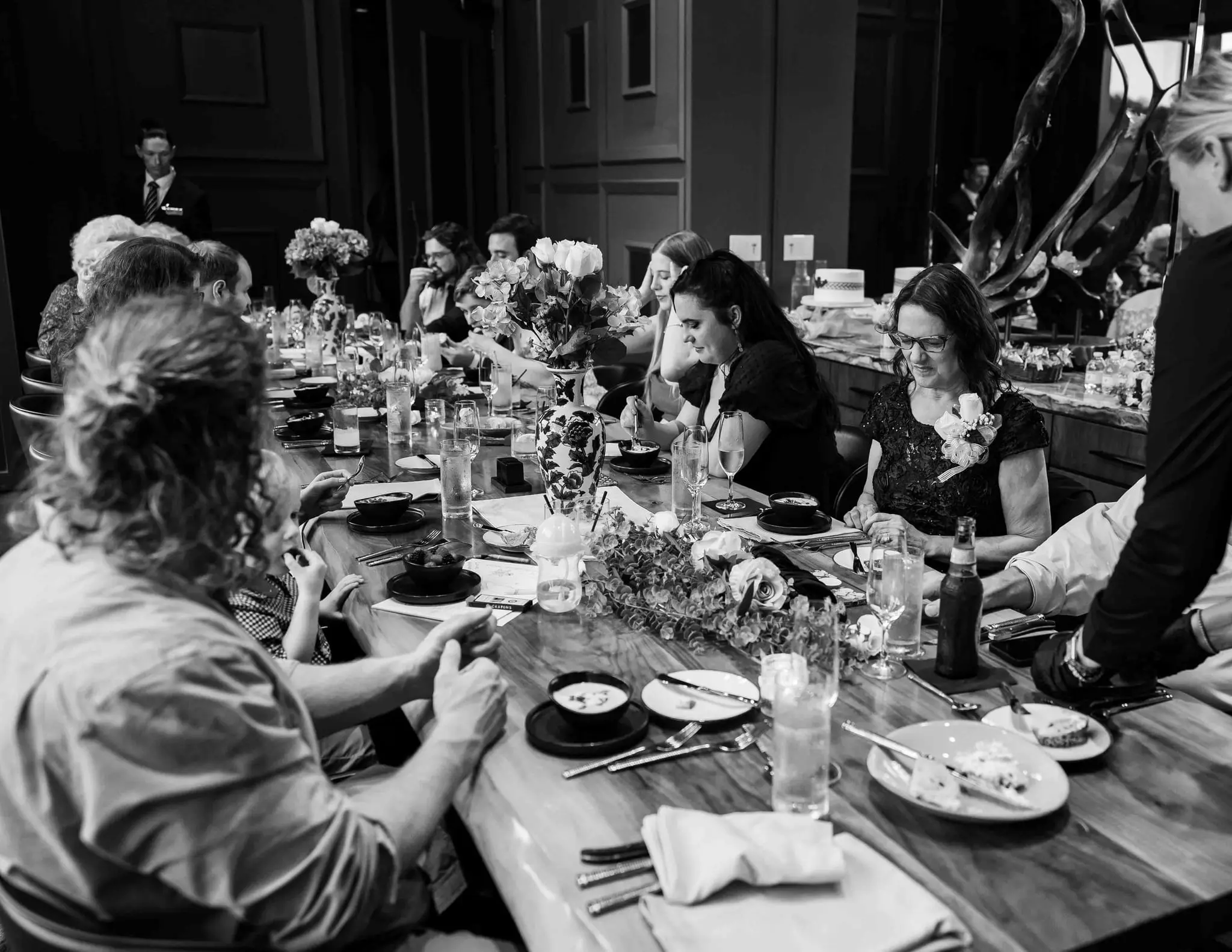 Black and white of guests dining along the long wooden reception table