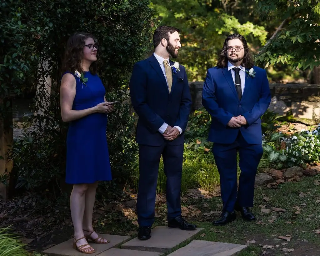 Attendant in a blue dress and two men in navy suits watching from the garden aisle