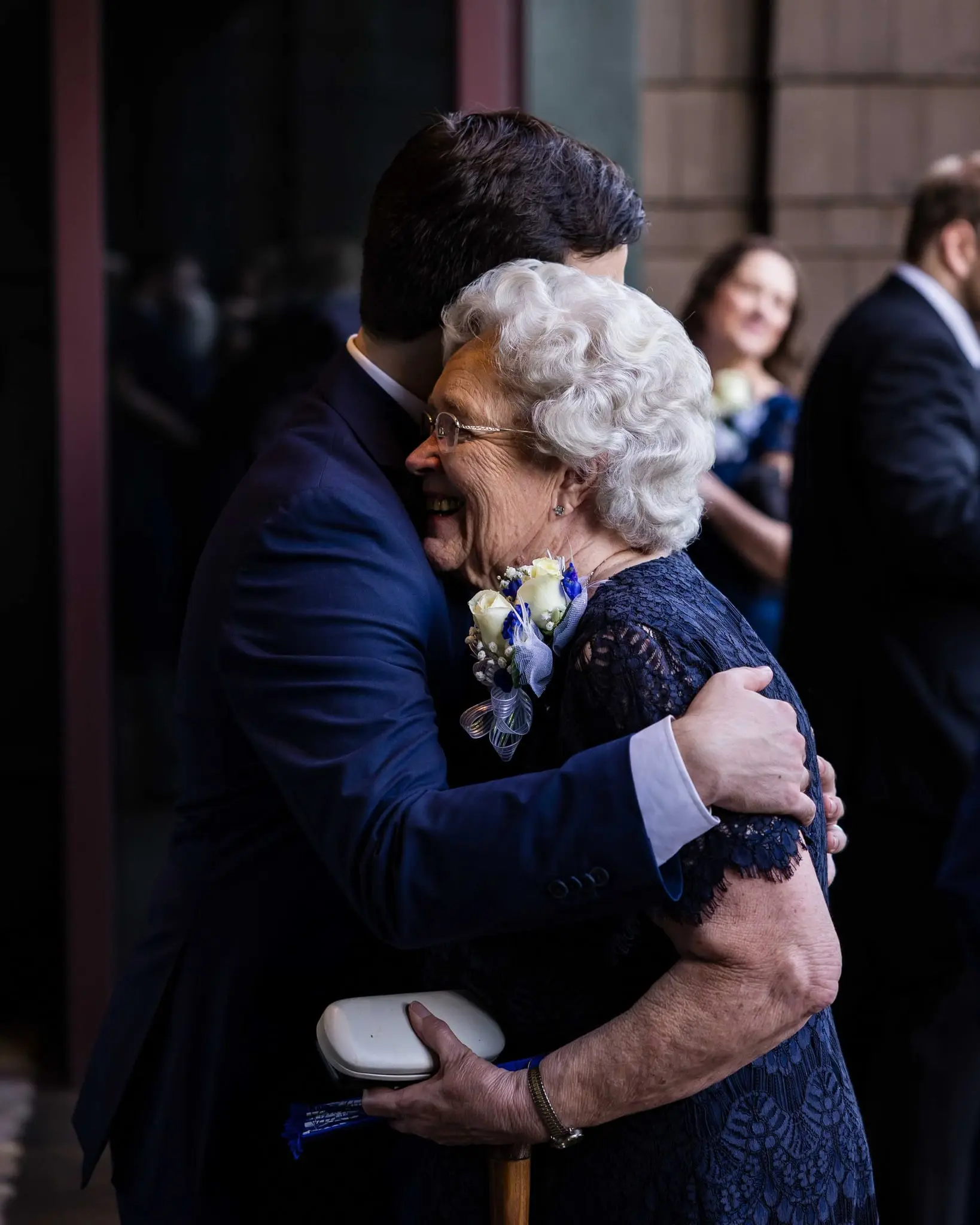 Groom in a navy suit hugging a smiling older woman with white hair and a flower corsage