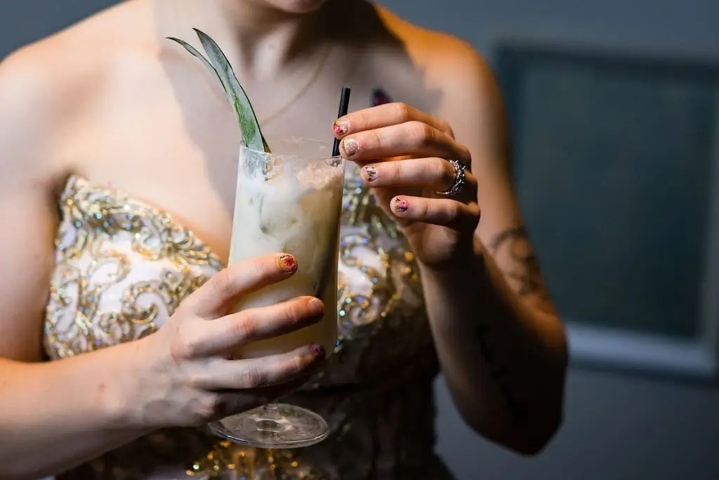 Bride holding a creamy cocktail garnished with a pineapple leaf, her ring and gold-beaded bodice visible