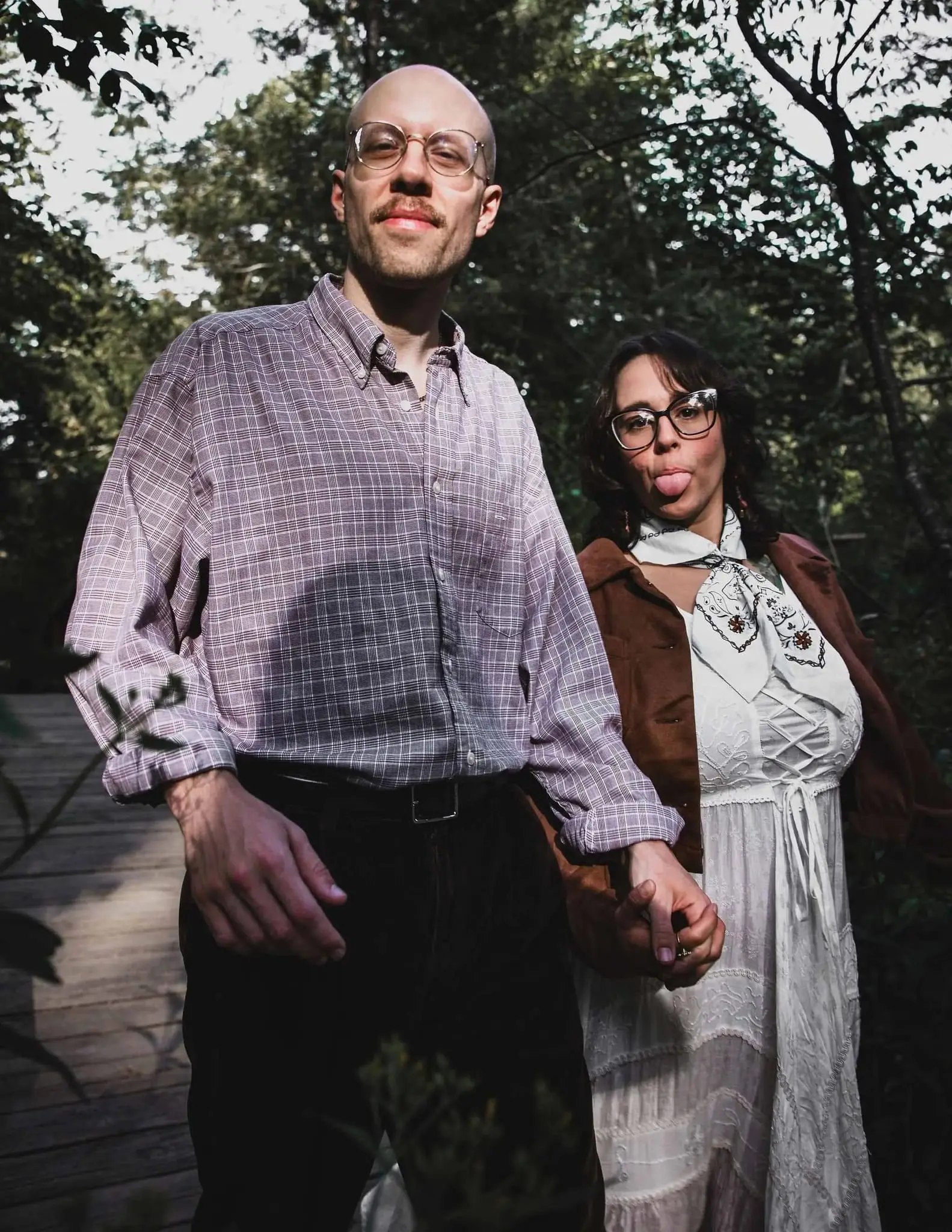 A man and woman standing outdoors on a wooden walkway, holding hands, surrounded by trees. The man is wearing a plaid shirt and glasses, and the woman is wearing a white dress with a neck scarf, glasses, and has her tongue sticking out.