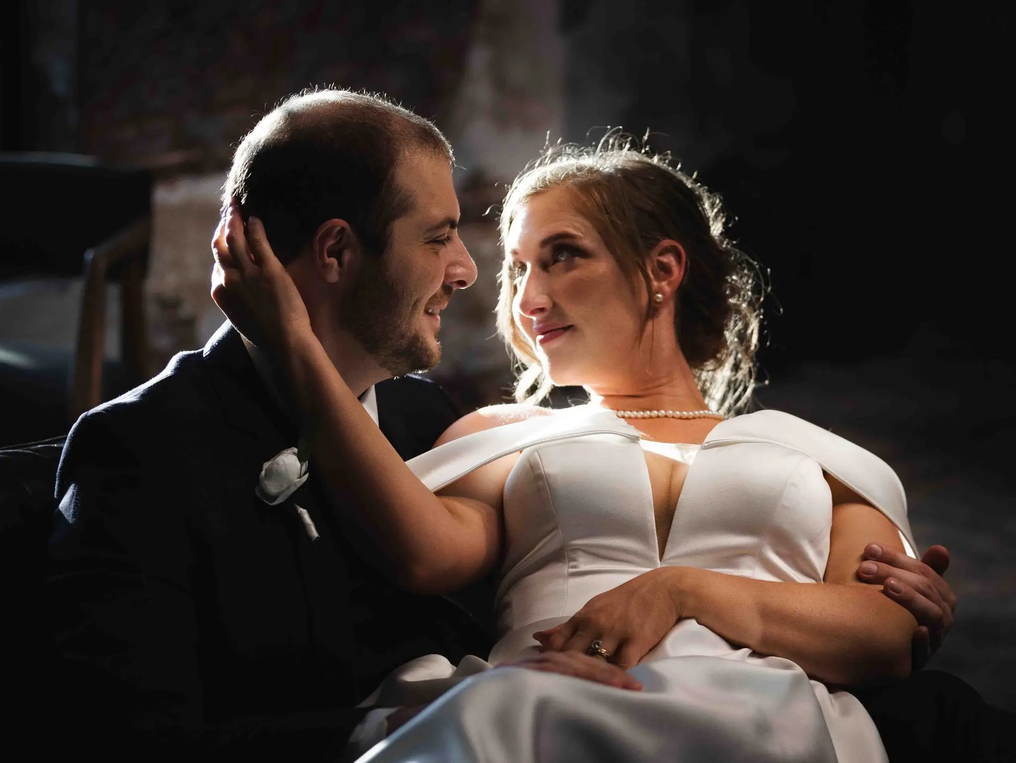 Bride and groom gazing at each other in dramatic window light, her pearl necklace and off-shoulder gown