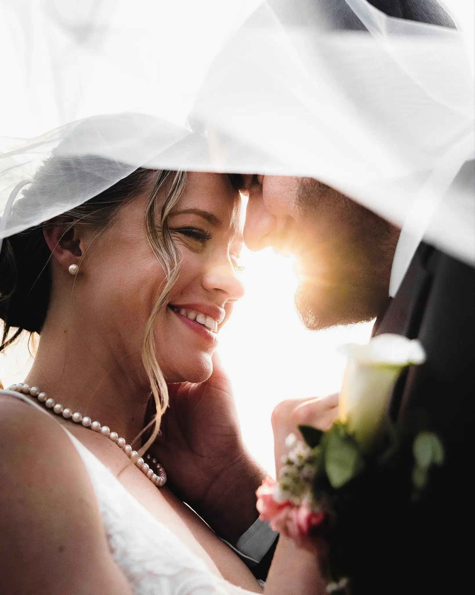 Bride and groom smiling forehead to forehead beneath her veil in backlit sun, a pearl necklace at her neck