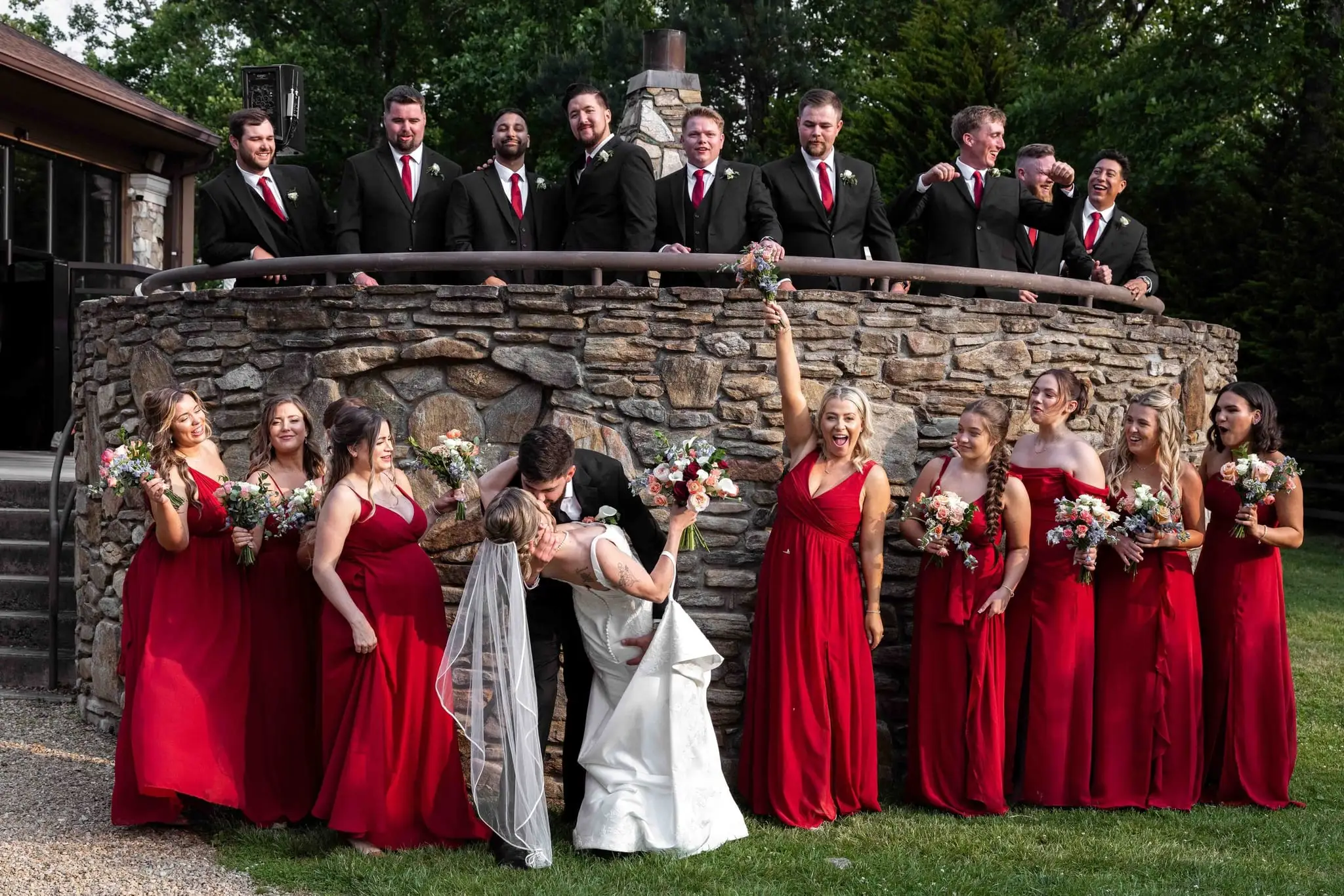 Anna and Joseph wedding at The Crest Center & Pavilion, Asheville NC — the bride in a white gown with bridesmaids in red dresses on a curved stone wall and groomsmen standing behind