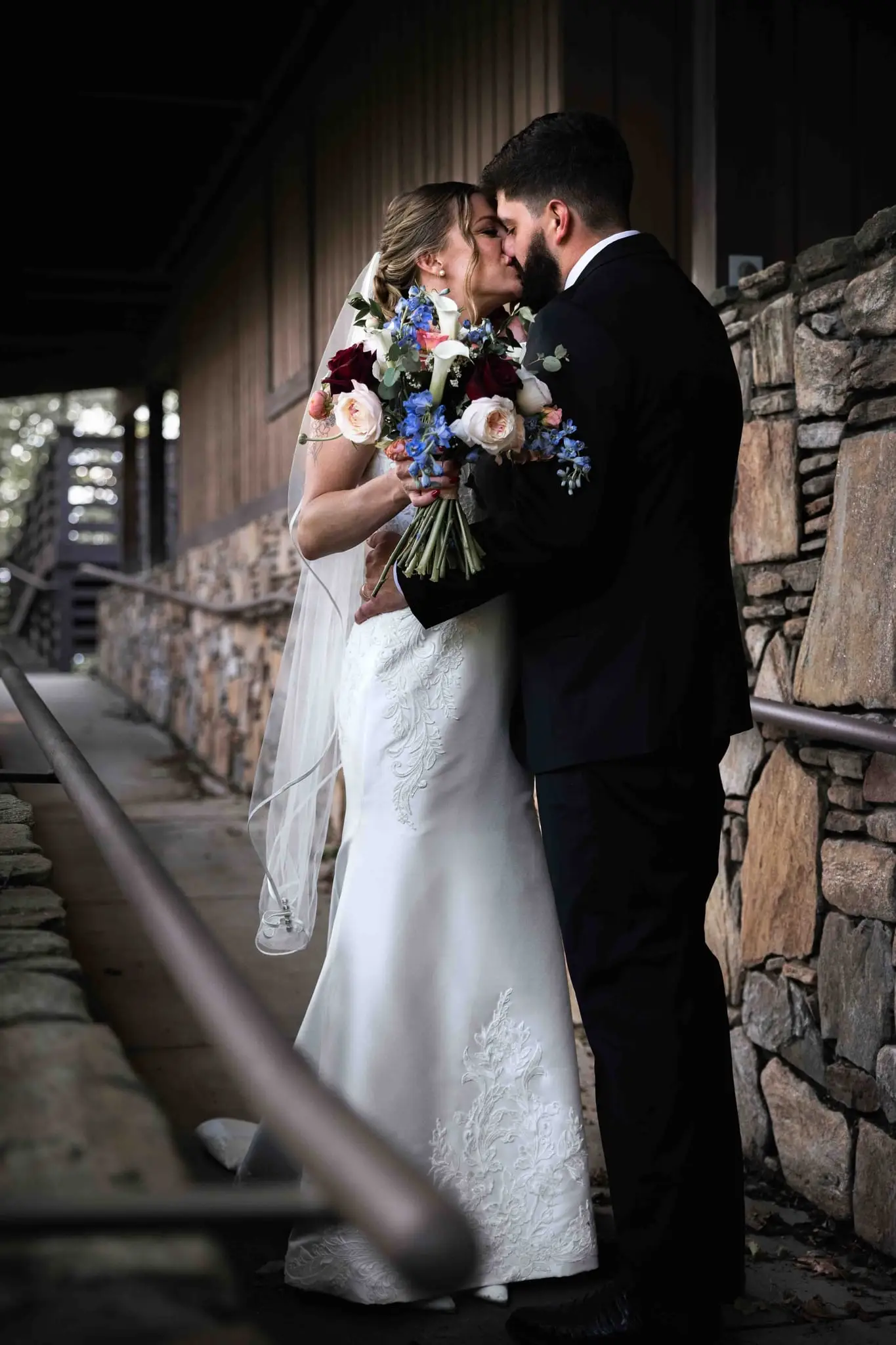 Anna and Joseph wedding at The Crest Center & Pavilion, Asheville NC — the couple kissing beside a stone wall and wooden railing, the bride holding a bouquet