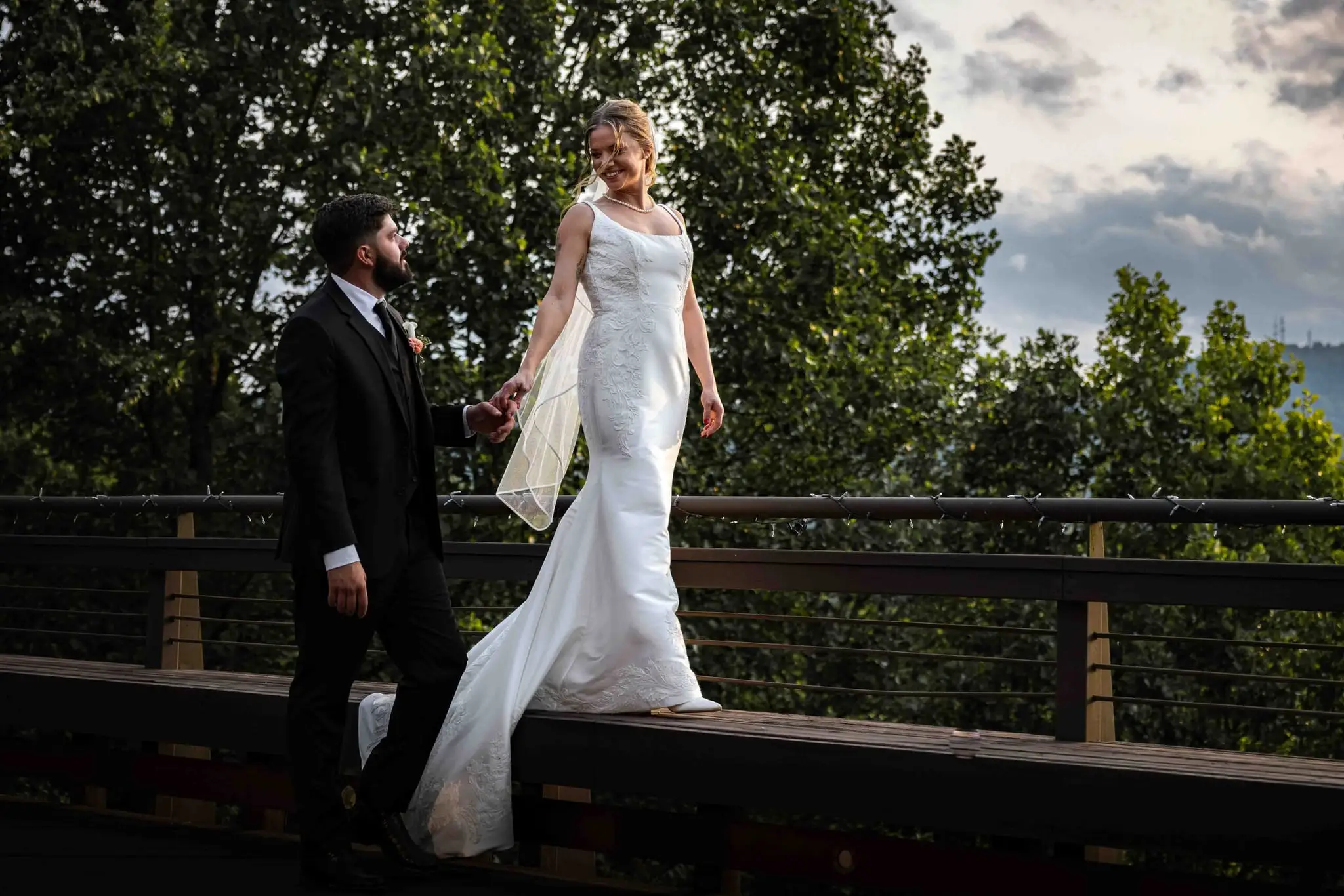 A bride walking along a deck rail above the mountains at the Crest Center in Asheville as her groom watches