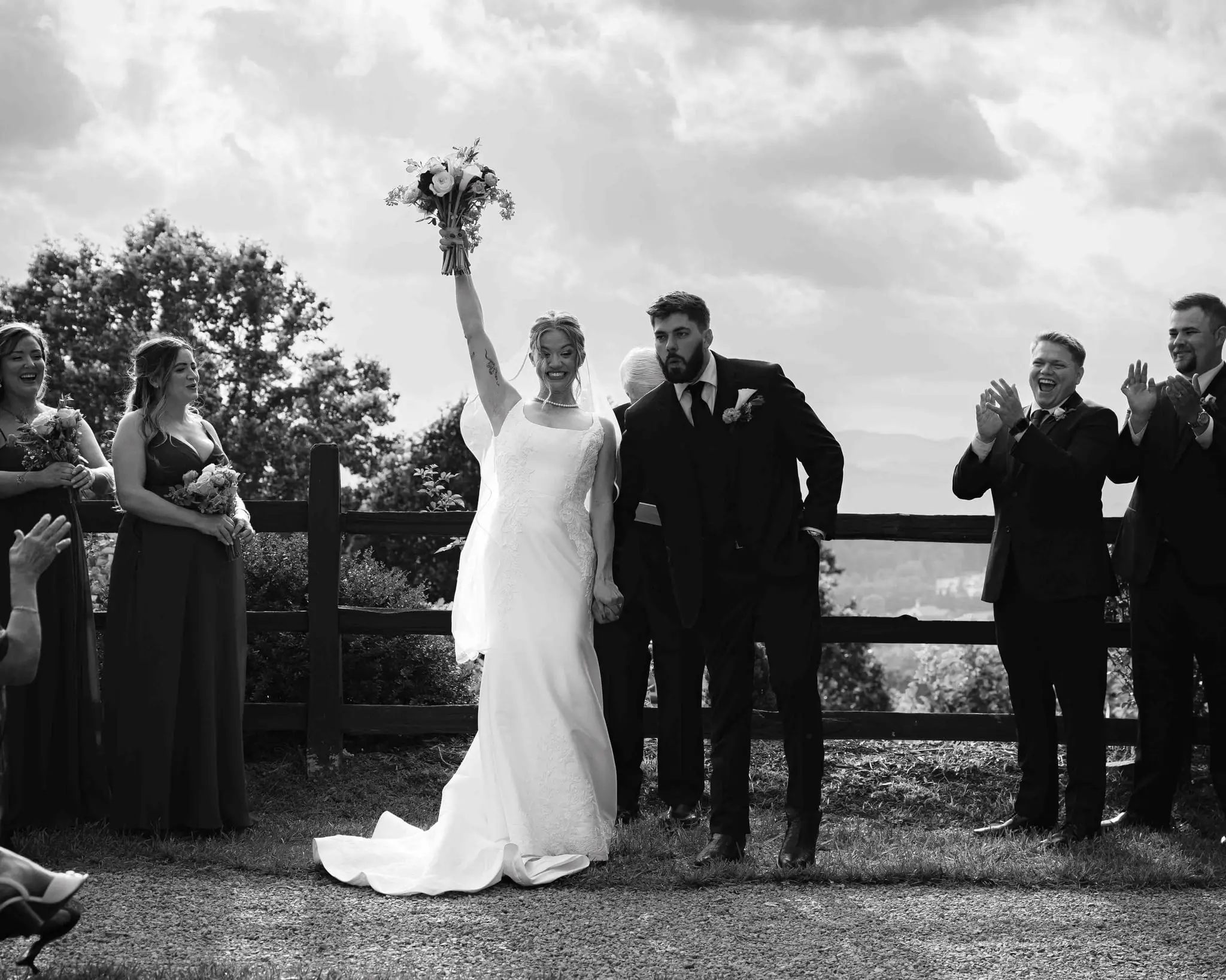 Anna and Joseph wedding at The Crest Center & Pavilion, Asheville NC — the bride raising her bouquet in celebration beside the groom at a wooden fence, black and white