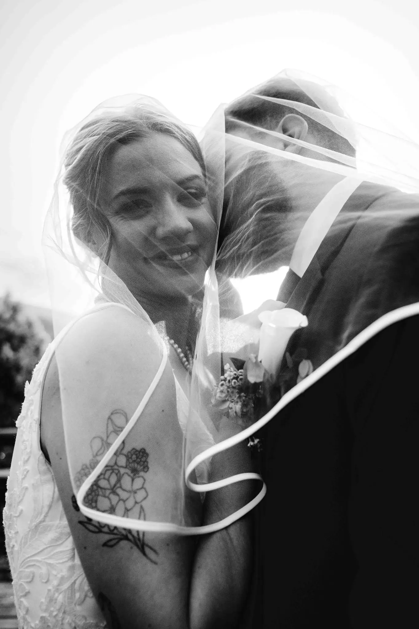 Black and white of a bride smiling through her veil as the groom nuzzles her, a floral tattoo on her shoulder