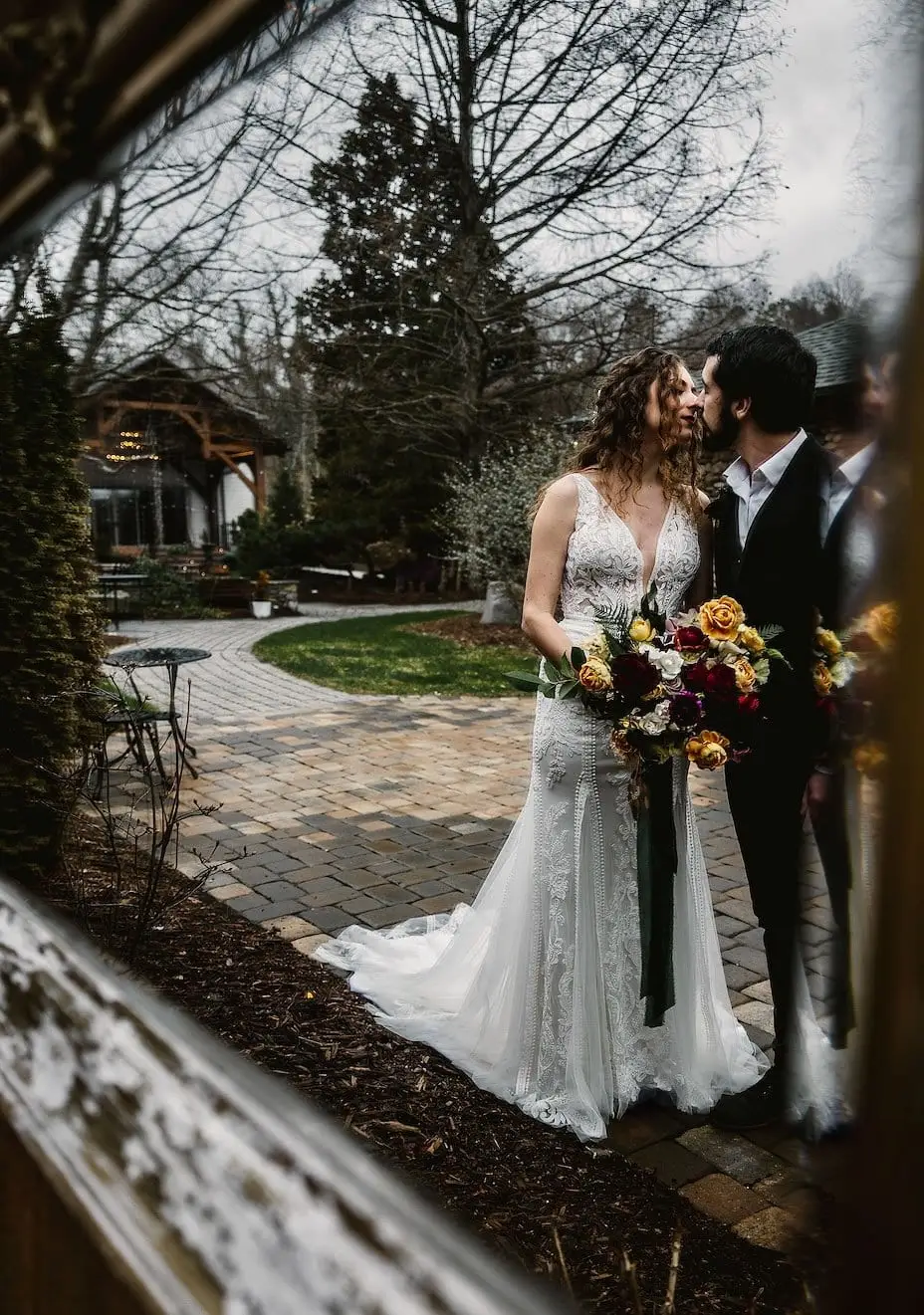 A bride and groom reflected in an ornate outdoor mirror among flowers at Haiku I Do