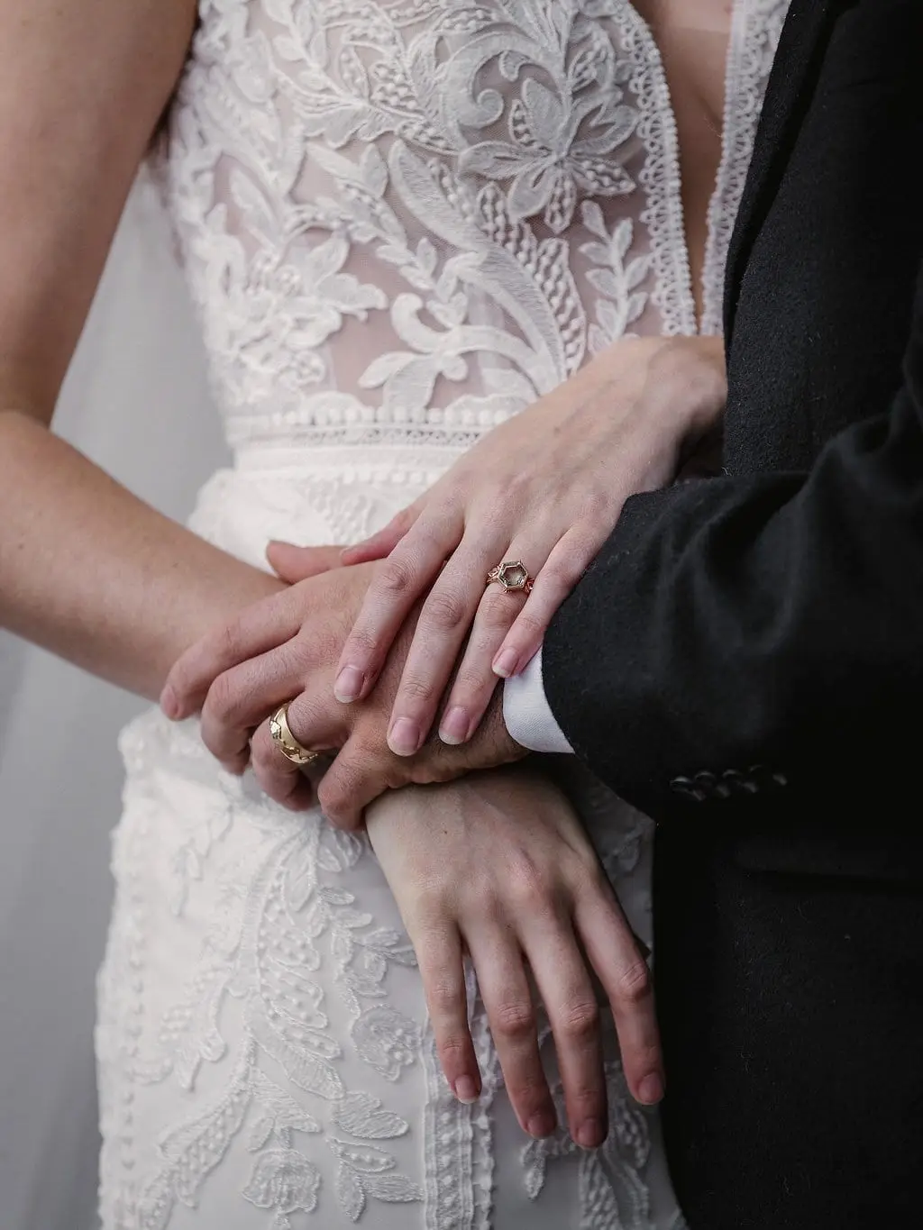 Close-up of the couple's hands and wedding rings against the bride's lace gown and the groom's dark suit