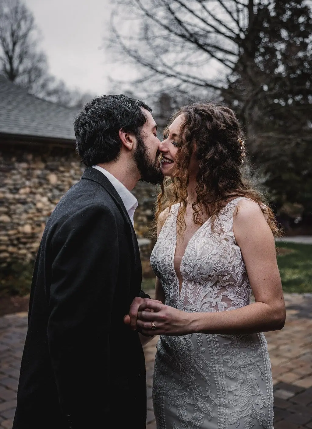 Groom leans in for a kiss on a brick path, the couple's hands clasped