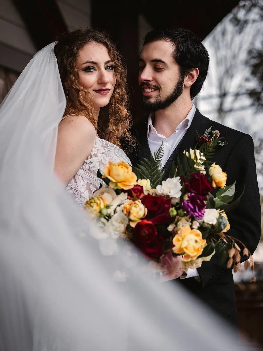 Smiling bride and groom pose close with a flowing veil and bouquet on a covered porch