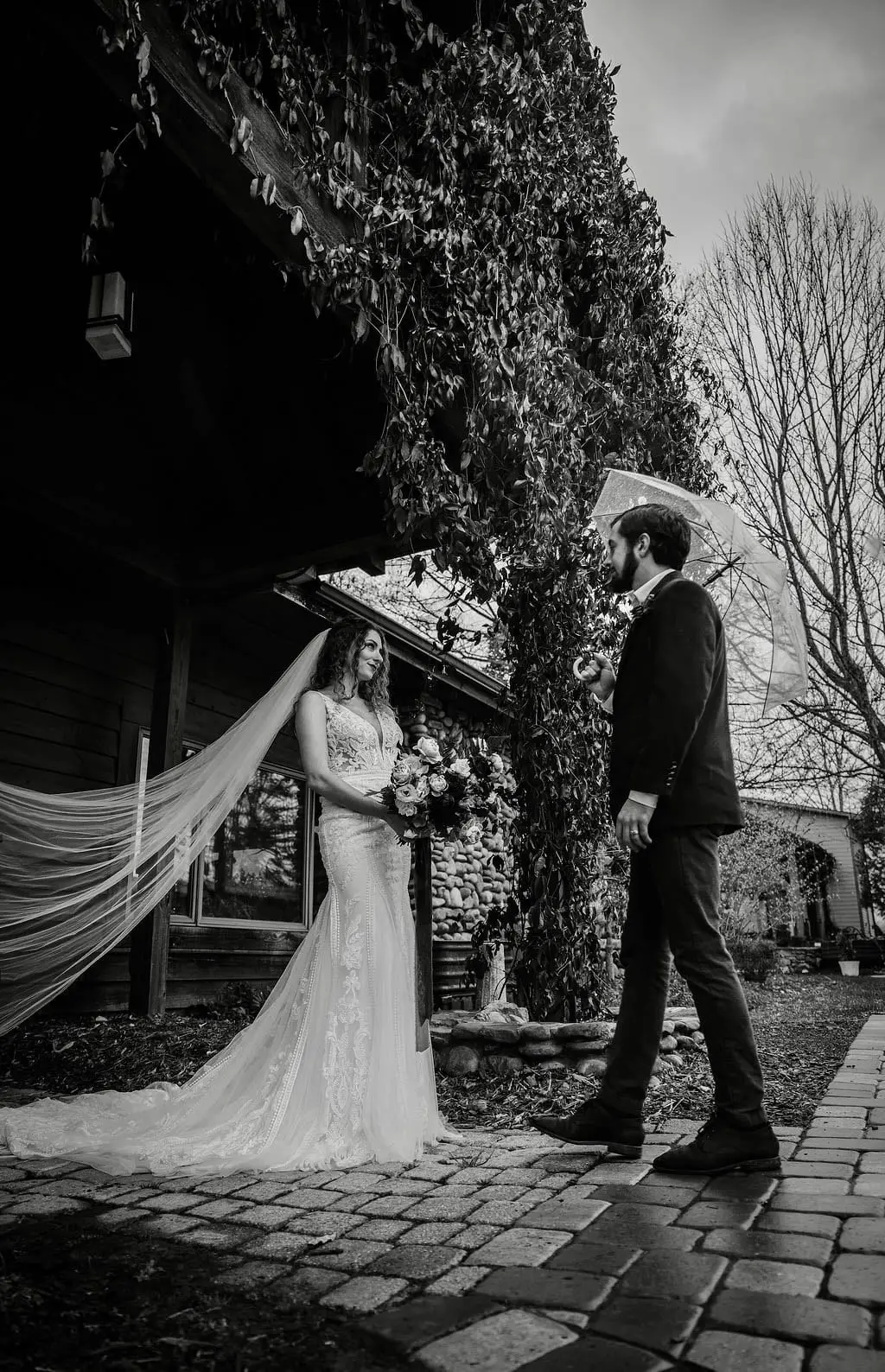 Black and white portrait of the couple under an ivy-covered porch, the bride's veil lifting in the wind