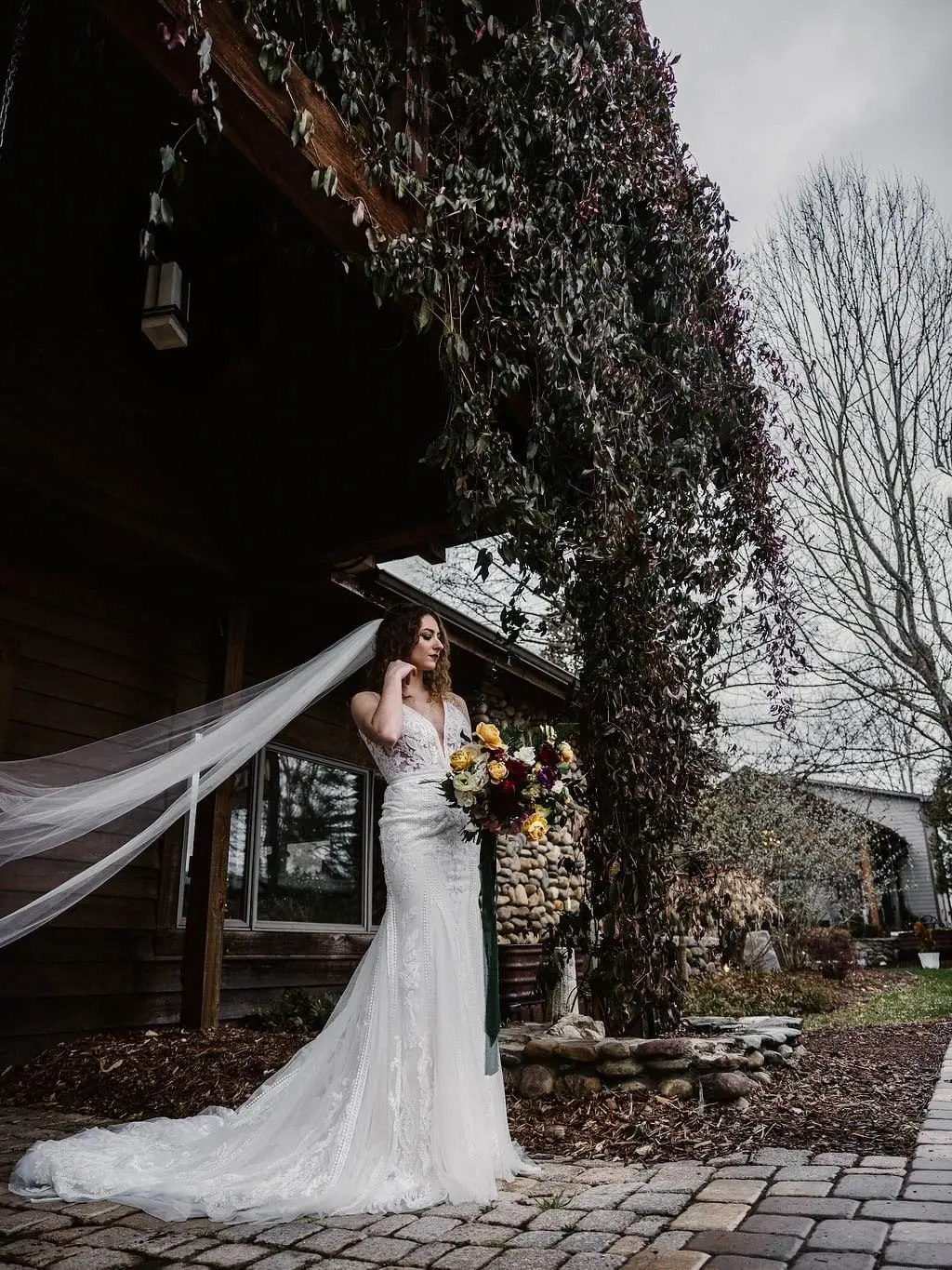 Bride holding her bouquet with a long veil blowing beside an ivy-draped timber lodge and bare winter trees