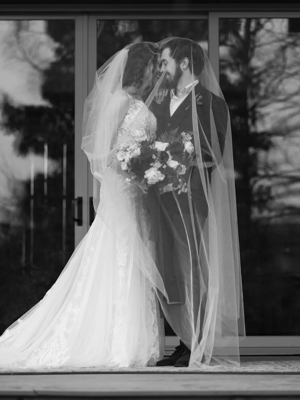 Black and white portrait of the couple sharing a veil on a deck in front of sliding glass doors