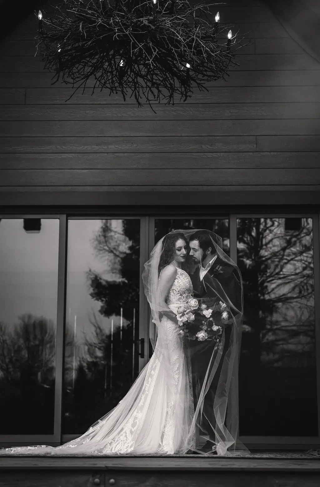 Black and white couple wrapped in a veil in front of glass doors below a branch chandelier
