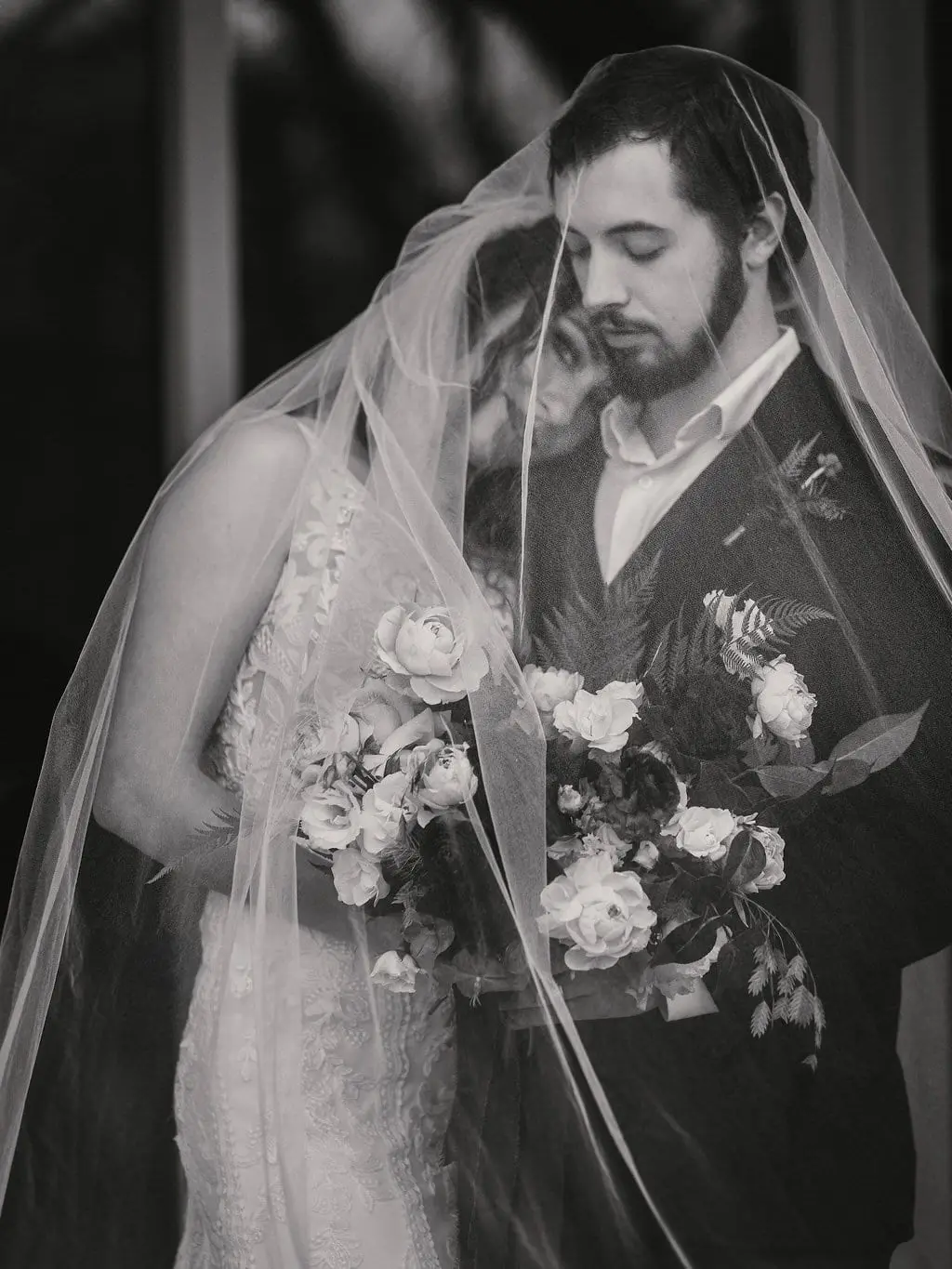 Black and white portrait of the couple under a sheer veil, the bride holding a white rose bouquet