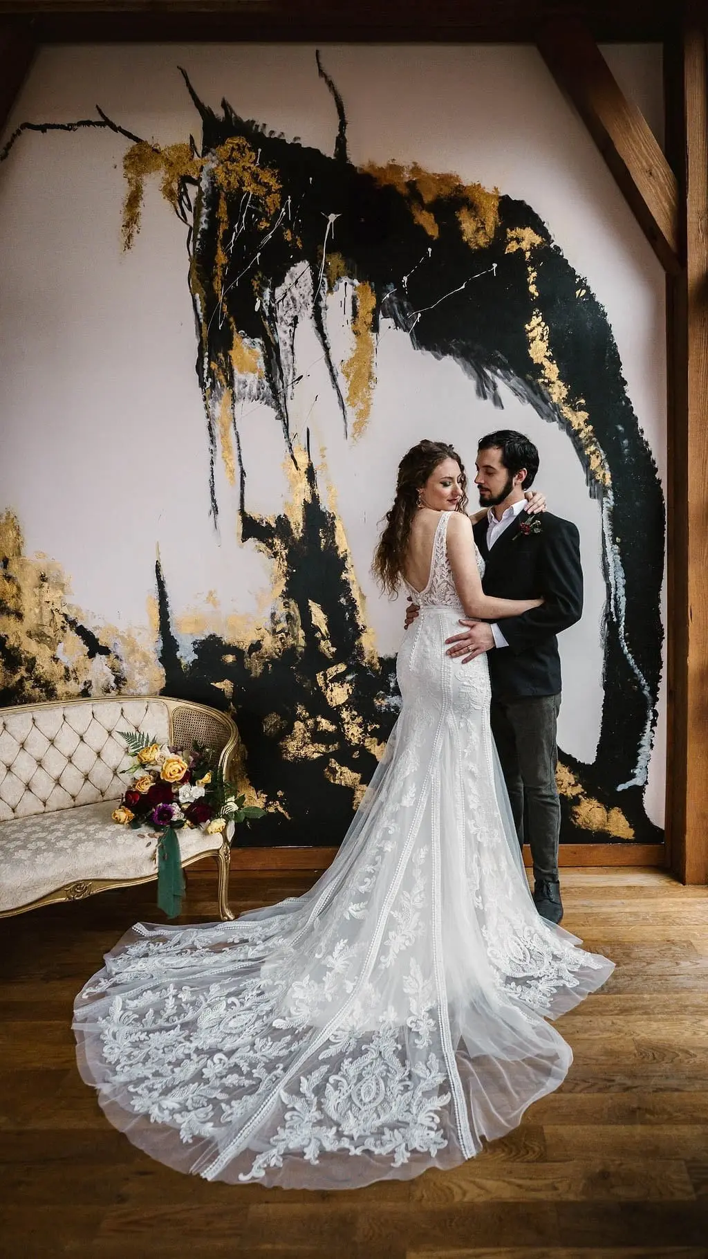 Couple embrace on a wood floor as the lace train of the bride's gown fans out before the painted wall