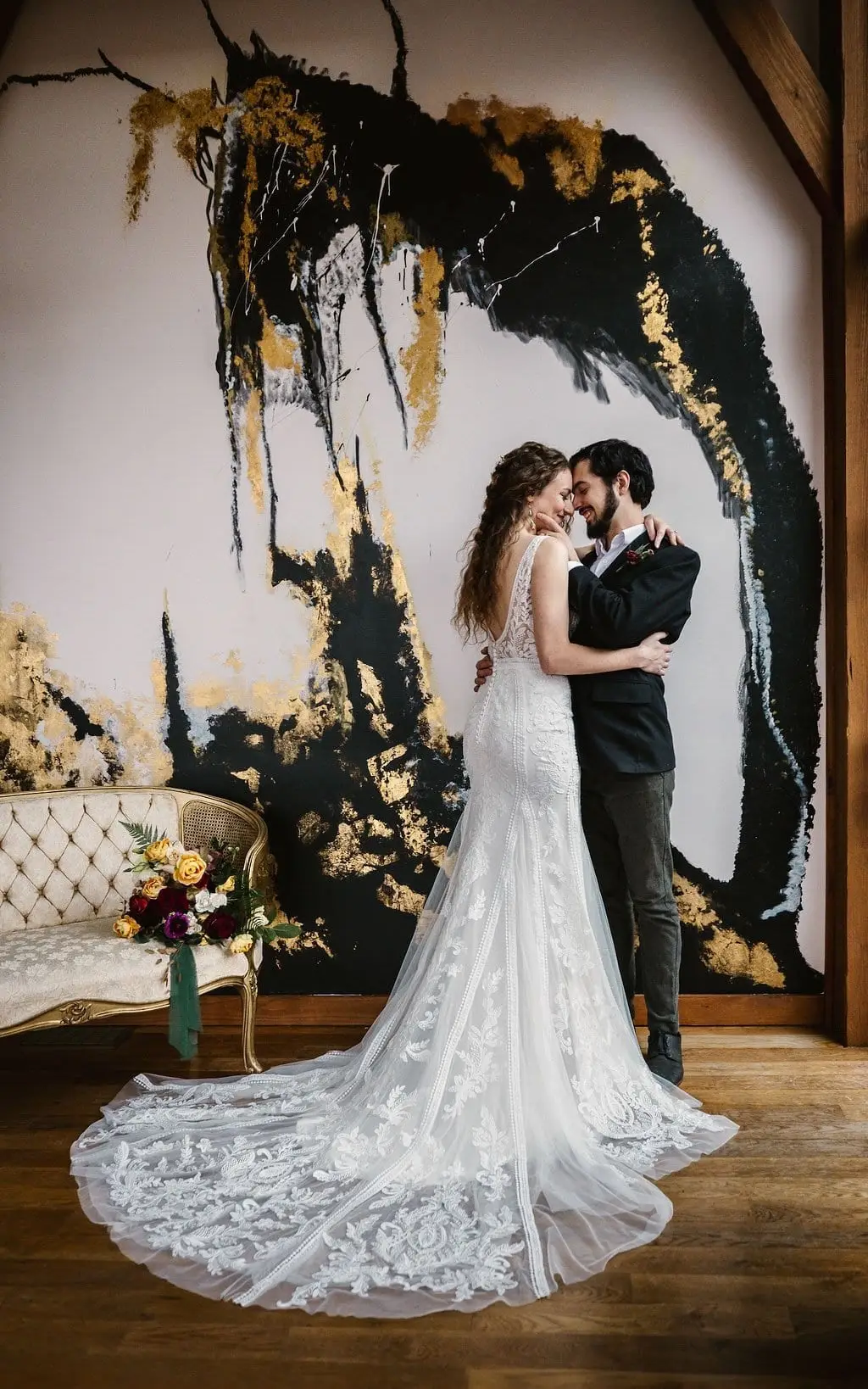 Couple dance close on a wood floor in front of the black and gold painted wall, the lace train spread behind