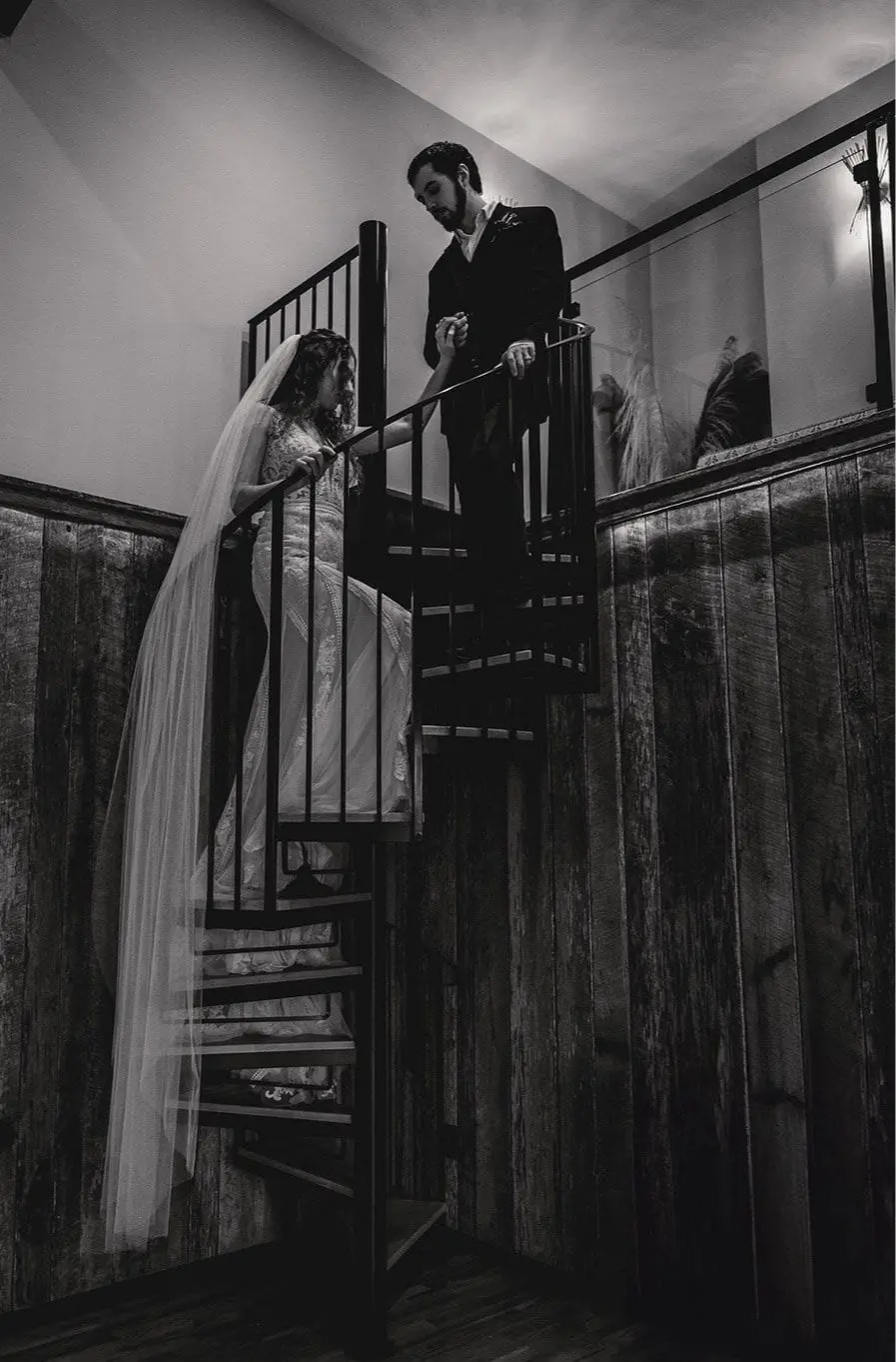 Black and white shot of the groom taking the bride's hand on a spiral staircase beneath a wood-paneled wall