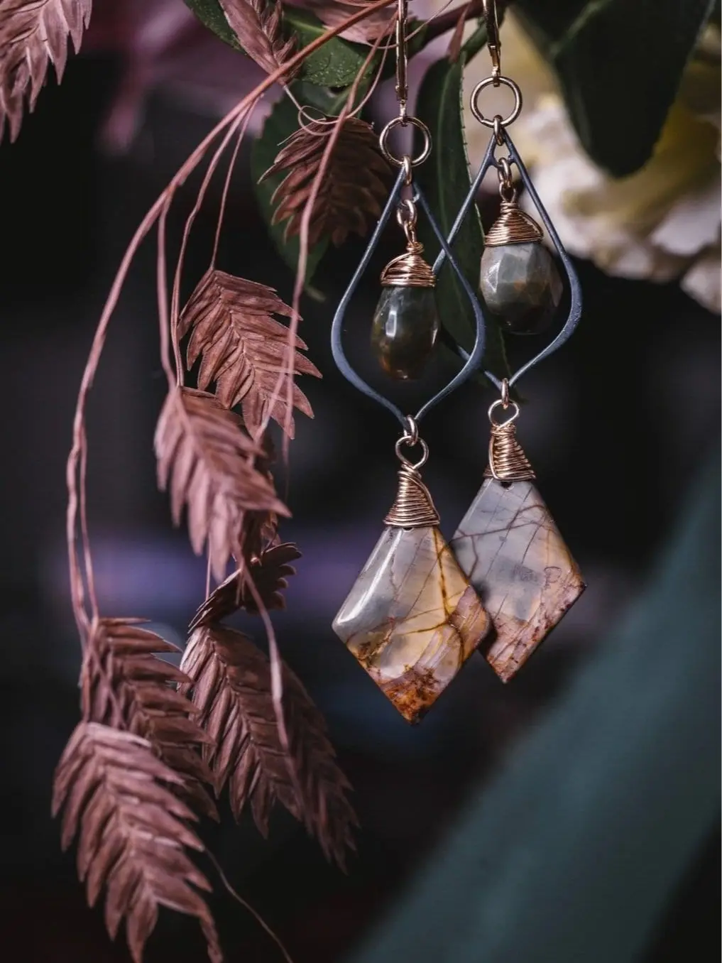 Wire-wrapped gemstone earrings hanging beside dried pink grass sprigs